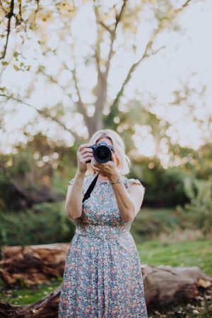 Photographer in floral dress holding camera in outdoor nature setting.
