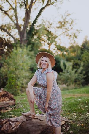 Person in floral dress and brown hat sitting on fallen log in sunlit garden.