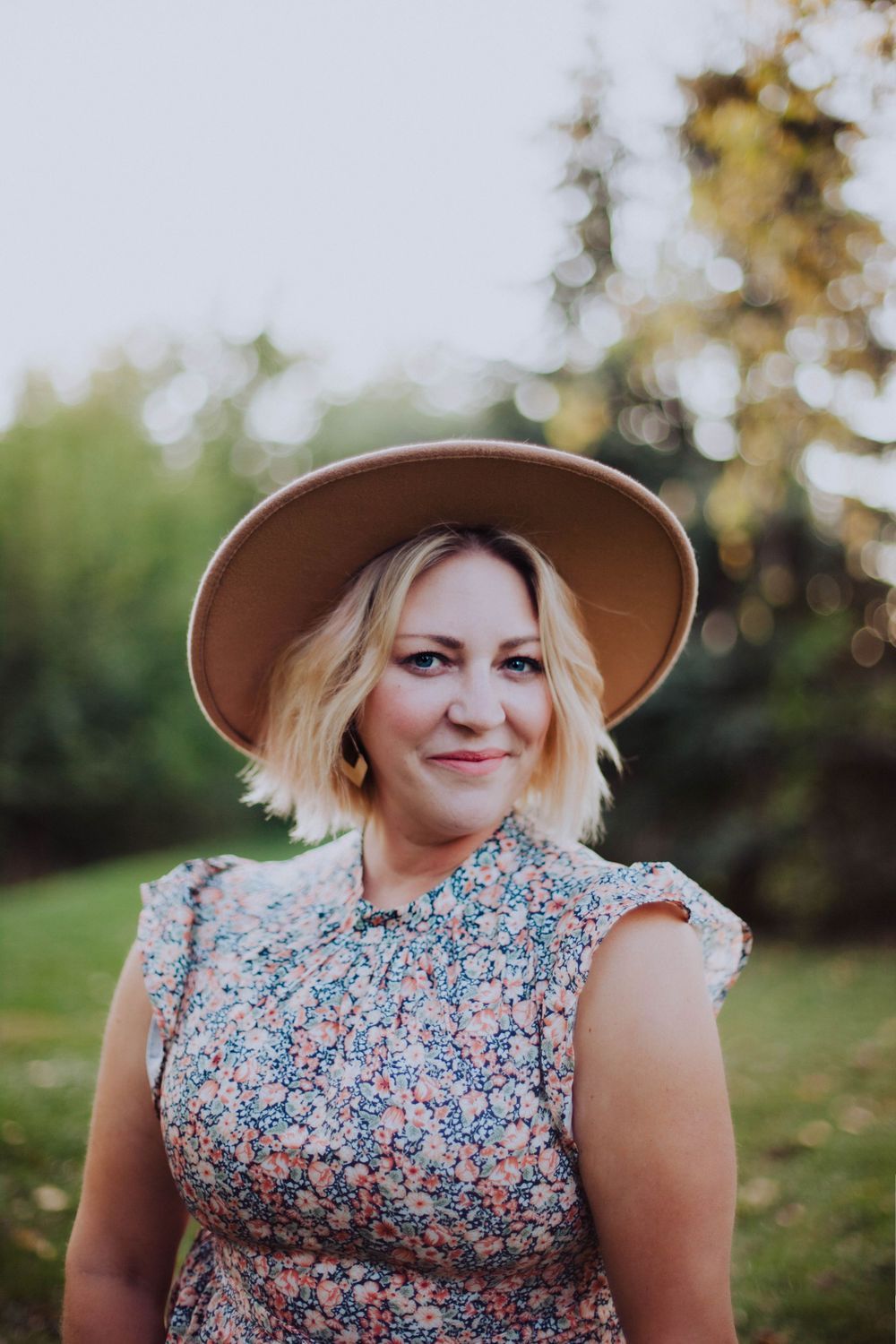 Natural light portrait of someone wearing brown hat and floral dress in garden setting.