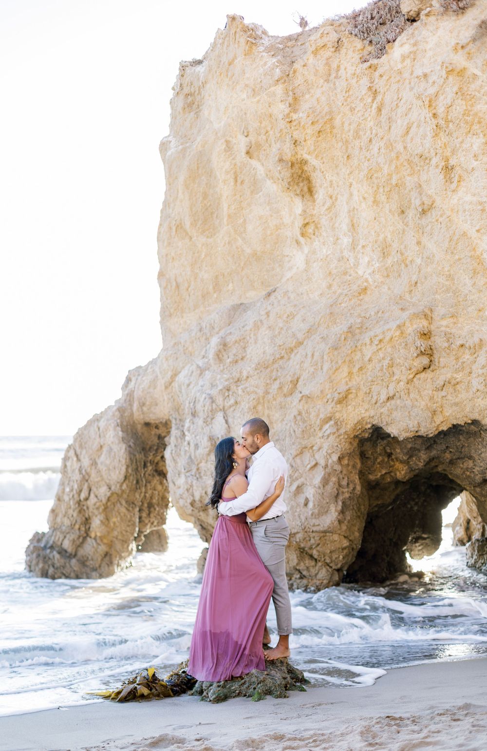 A romantic moment captured between a couple standing by a natural rock arch on a beach.