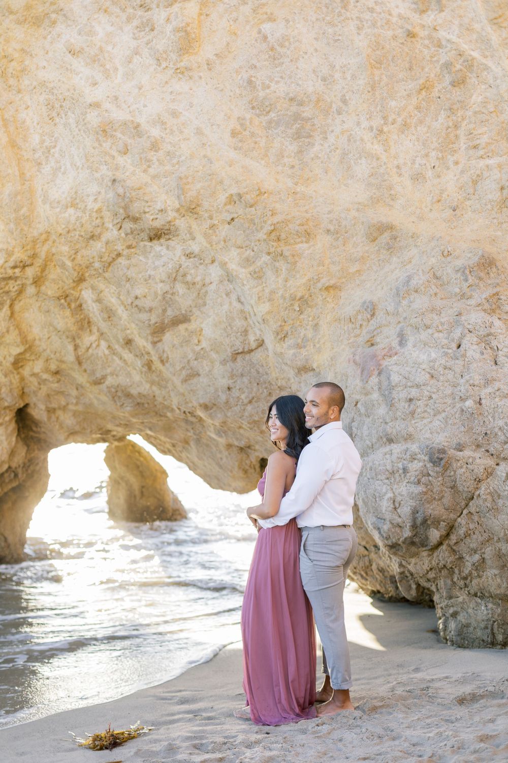 Romantic couple embracing on beach near limestone arch during golden hour.