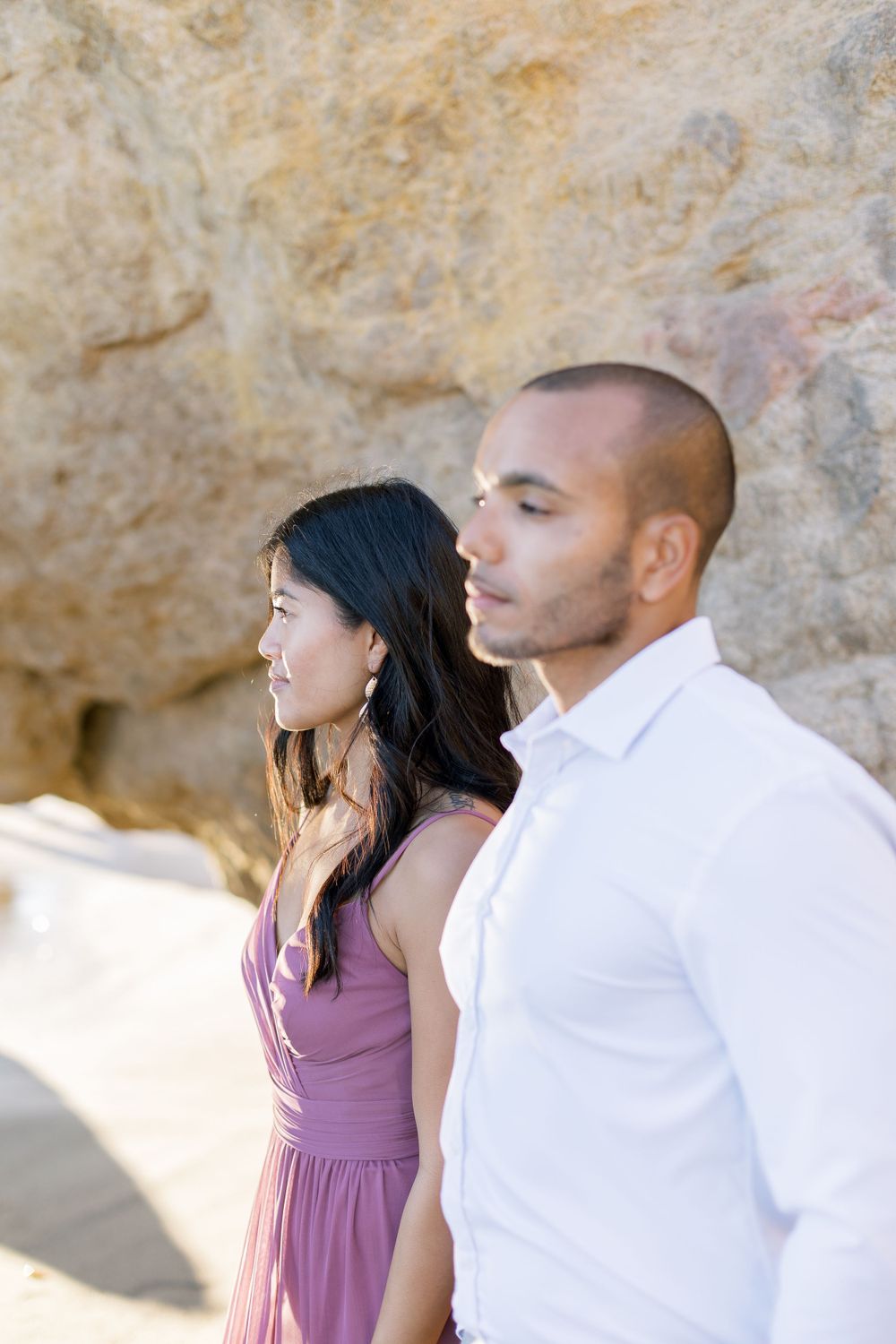 A couple in contrasting white and mauve outfits poses against a rocky beach backdrop.