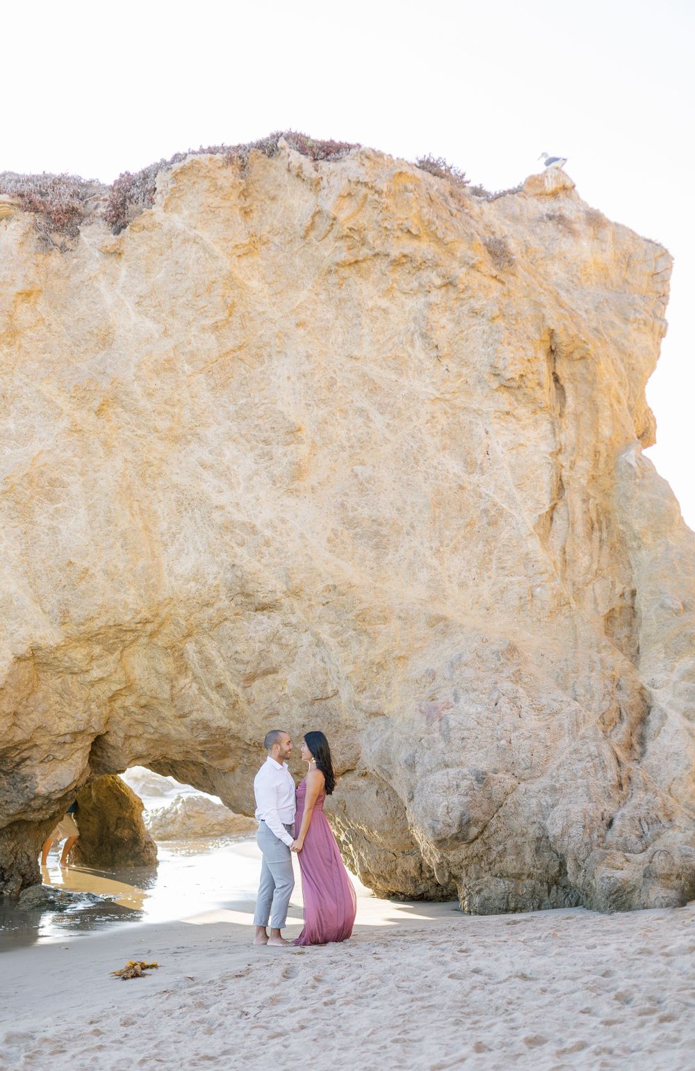 A couple stands beneath a dramatic limestone arch formation at the beach during golden hour.