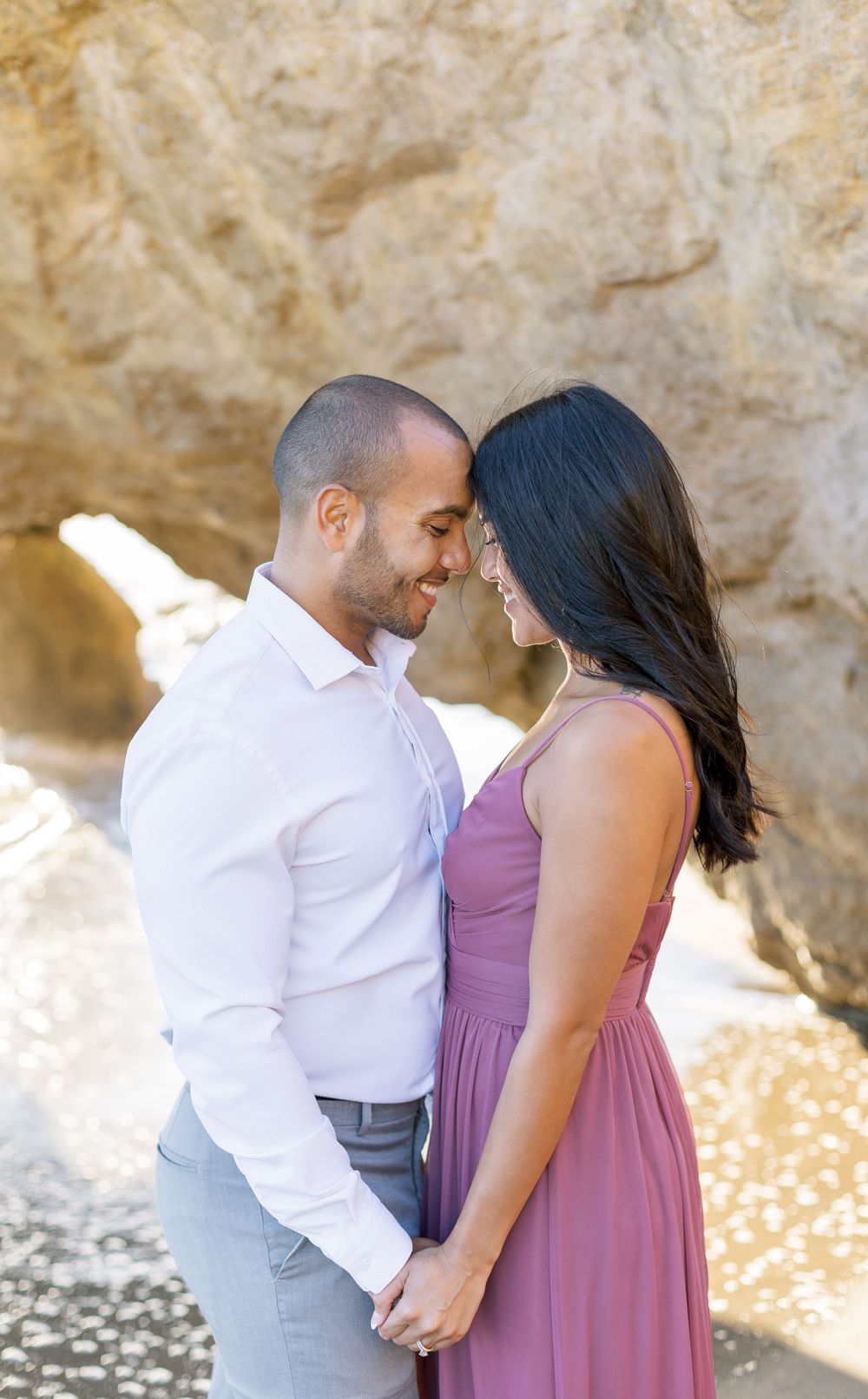 A couple shares a tender moment together near rocky cliffs at the beach.