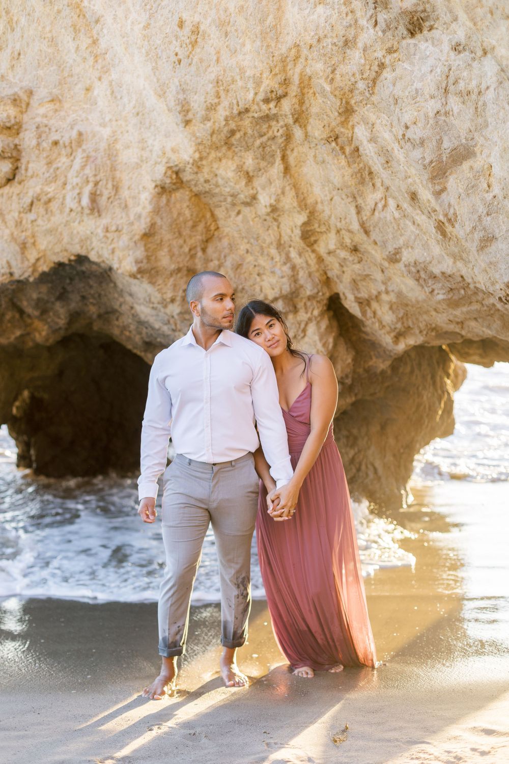 A couple holds hands while standing on a beach with dramatic sea caves behind them.