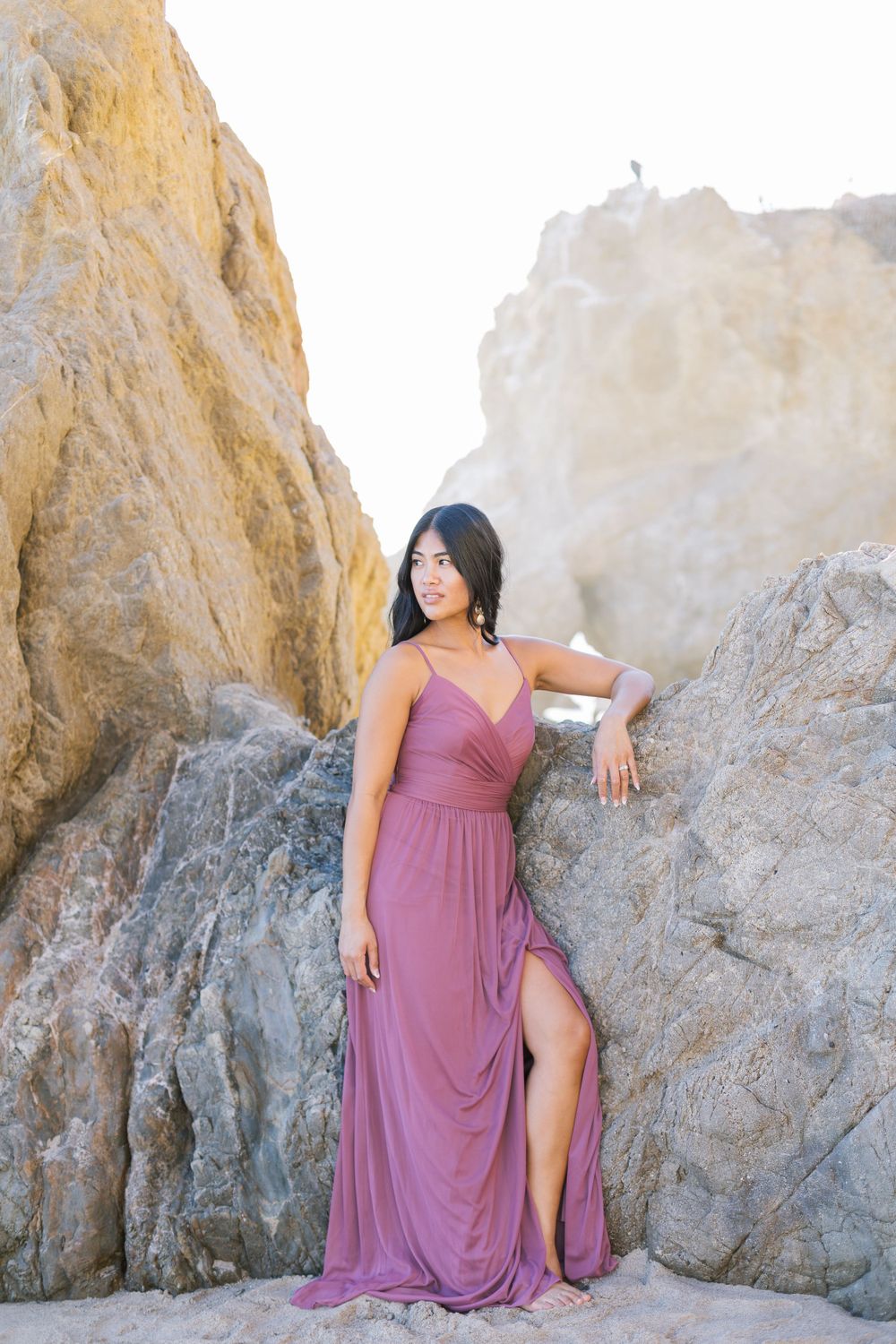 Woman in flowing mauve dress posing elegantly against coastal rock formations.