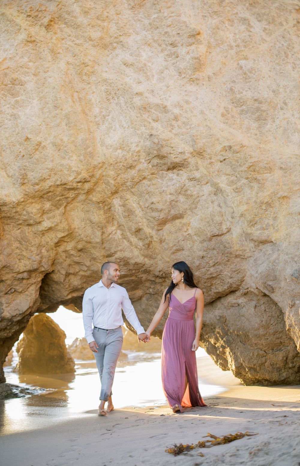 Couple holding hands while walking through natural rock arch at beach.