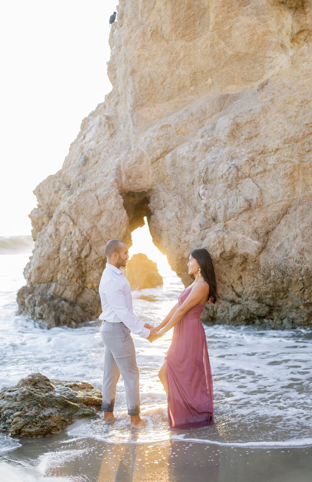 Couple standing in golden sunlight through rock arch at beach during sunset.