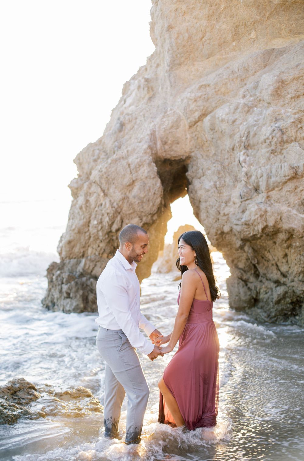 Couple in elegant outfits playing in ocean waves near rocky beach arch.