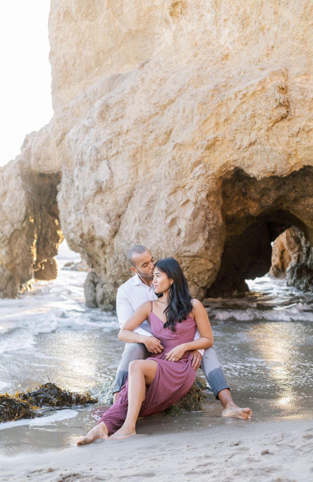 A couple embraces on a beach near dramatic limestone cliffs with natural sea caves at sunset.