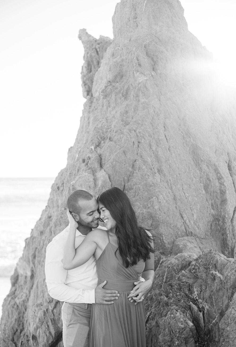 A black and white portrait of a couple sharing an intimate moment by coastal cliffs.