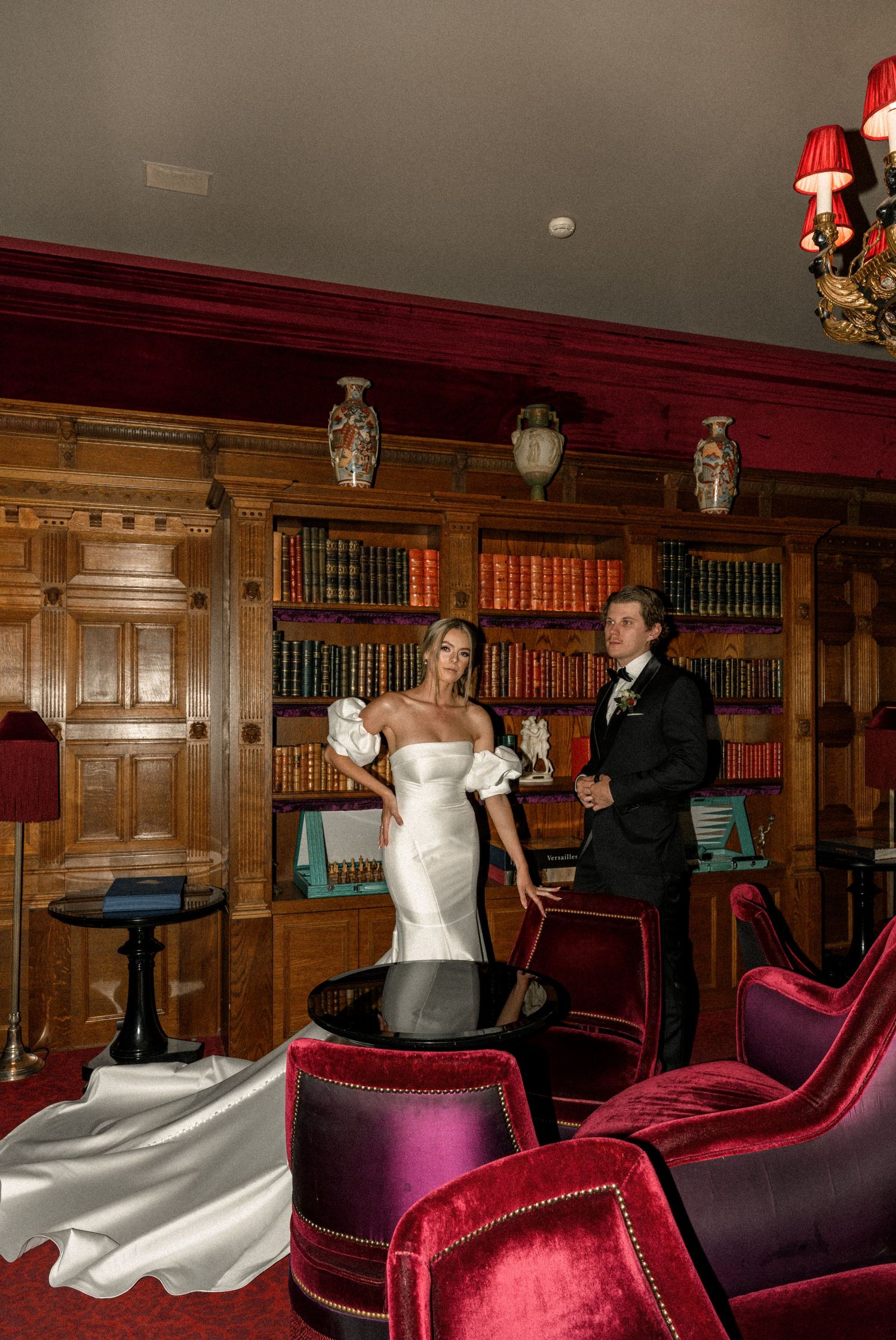 A wedding couple celebrates in an elegant wood-paneled library with plush burgundy velvet chairs.