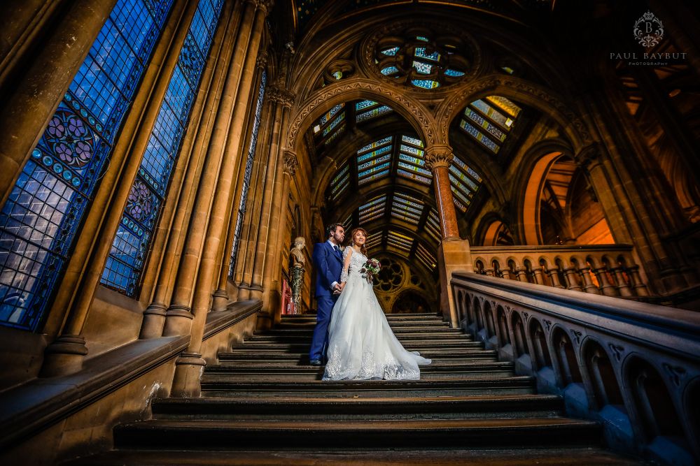 Couple in wedding attire pose on grand stone staircase with gothic architecture.