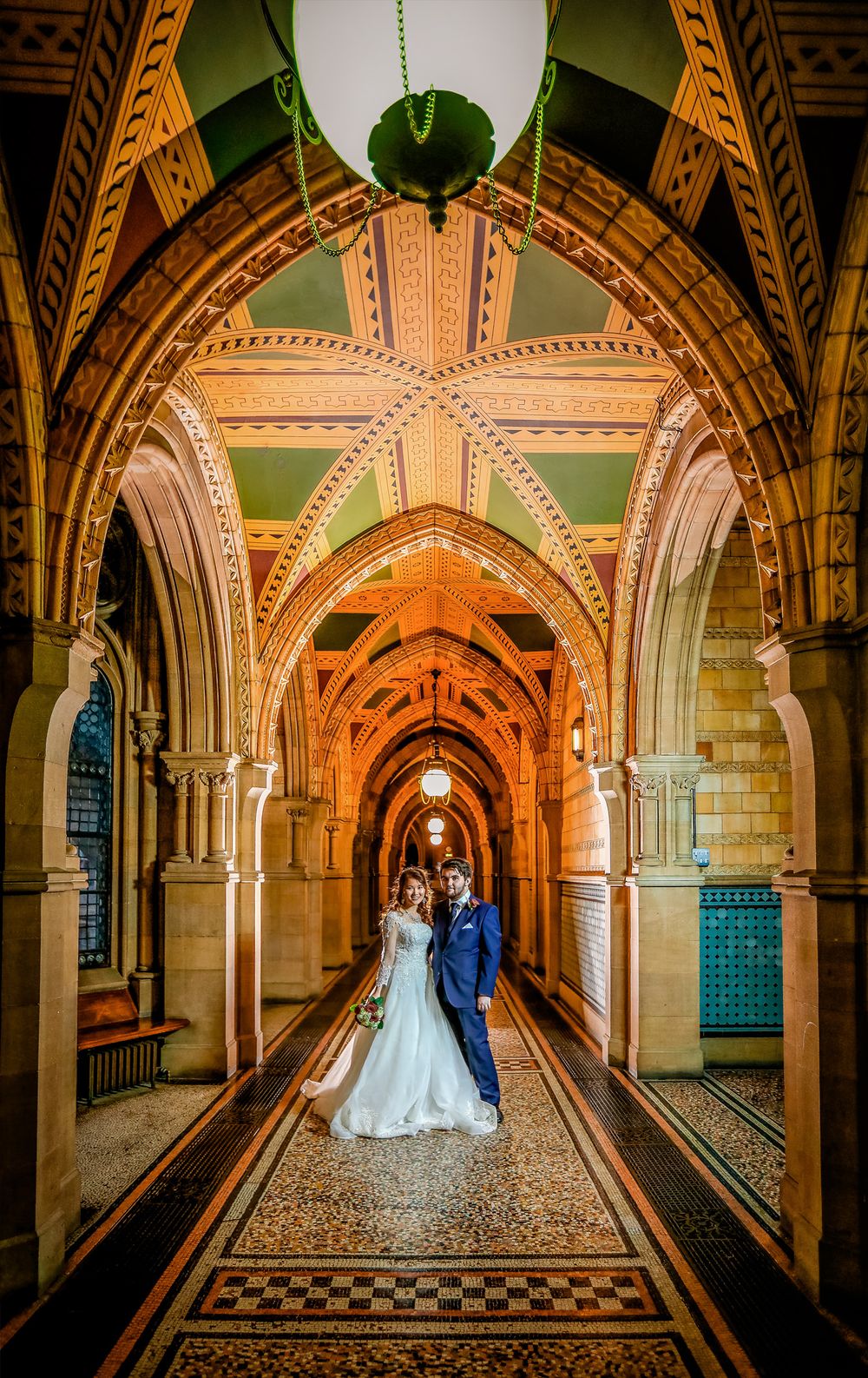 Newlyweds stand in ornate corridor with patterned ceiling and arches.