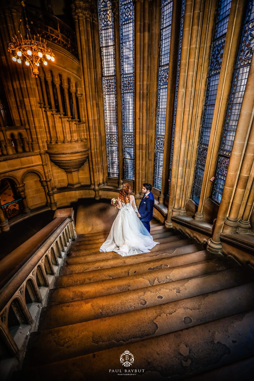 Newlyweds stand on ornate staircase with dramatic stained glass windows.
