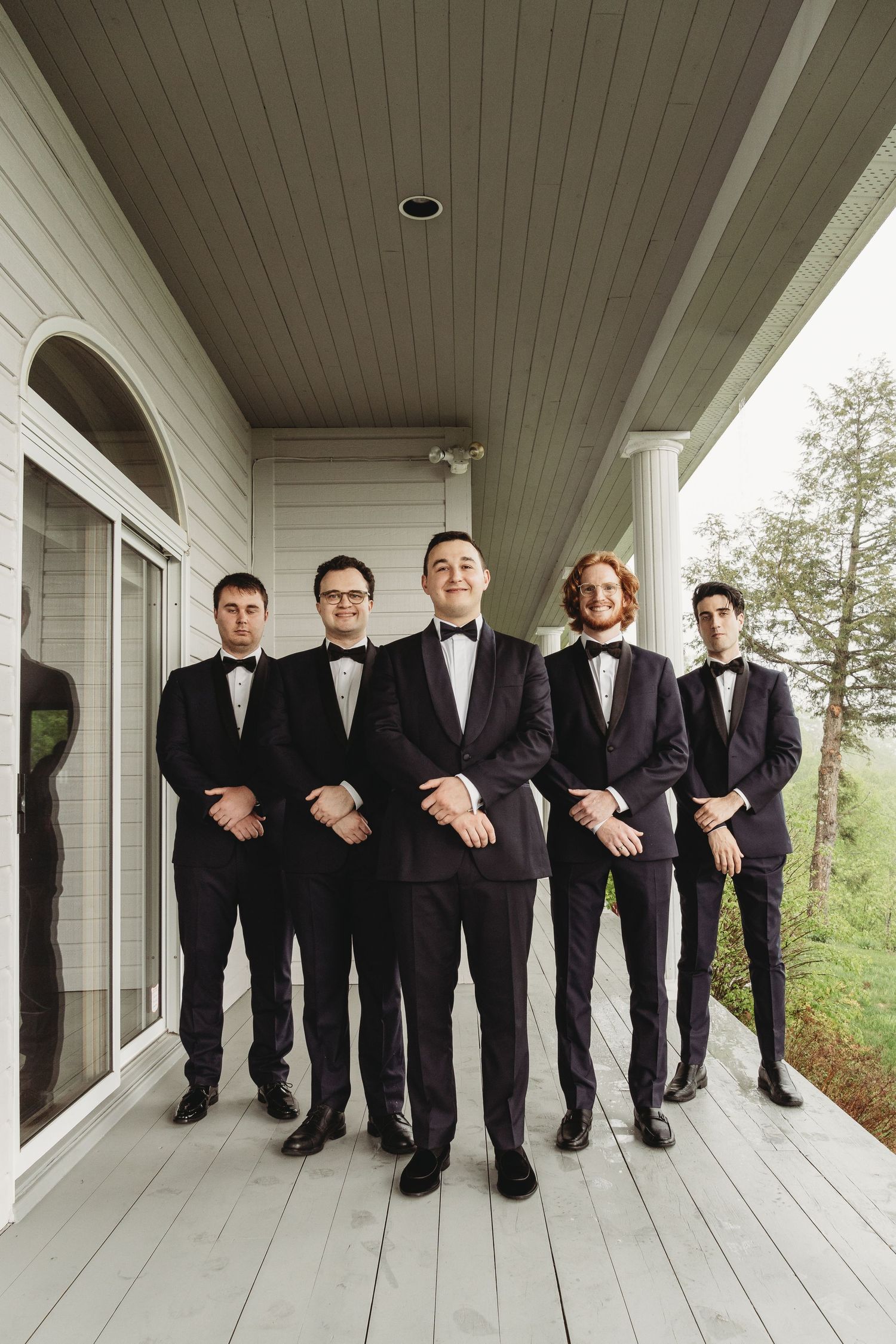 Five people in black tuxedos and bow ties lined up on a white porch for a formal photo.