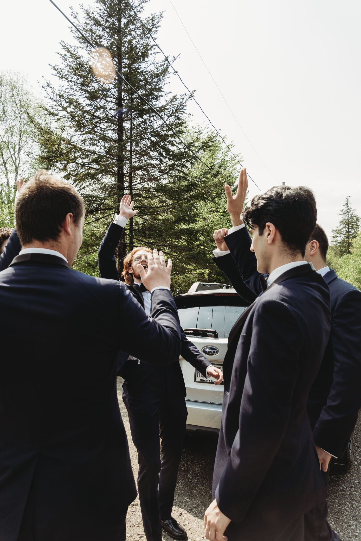 Group in dark suits raising their hands in celebration outdoors near pine trees.