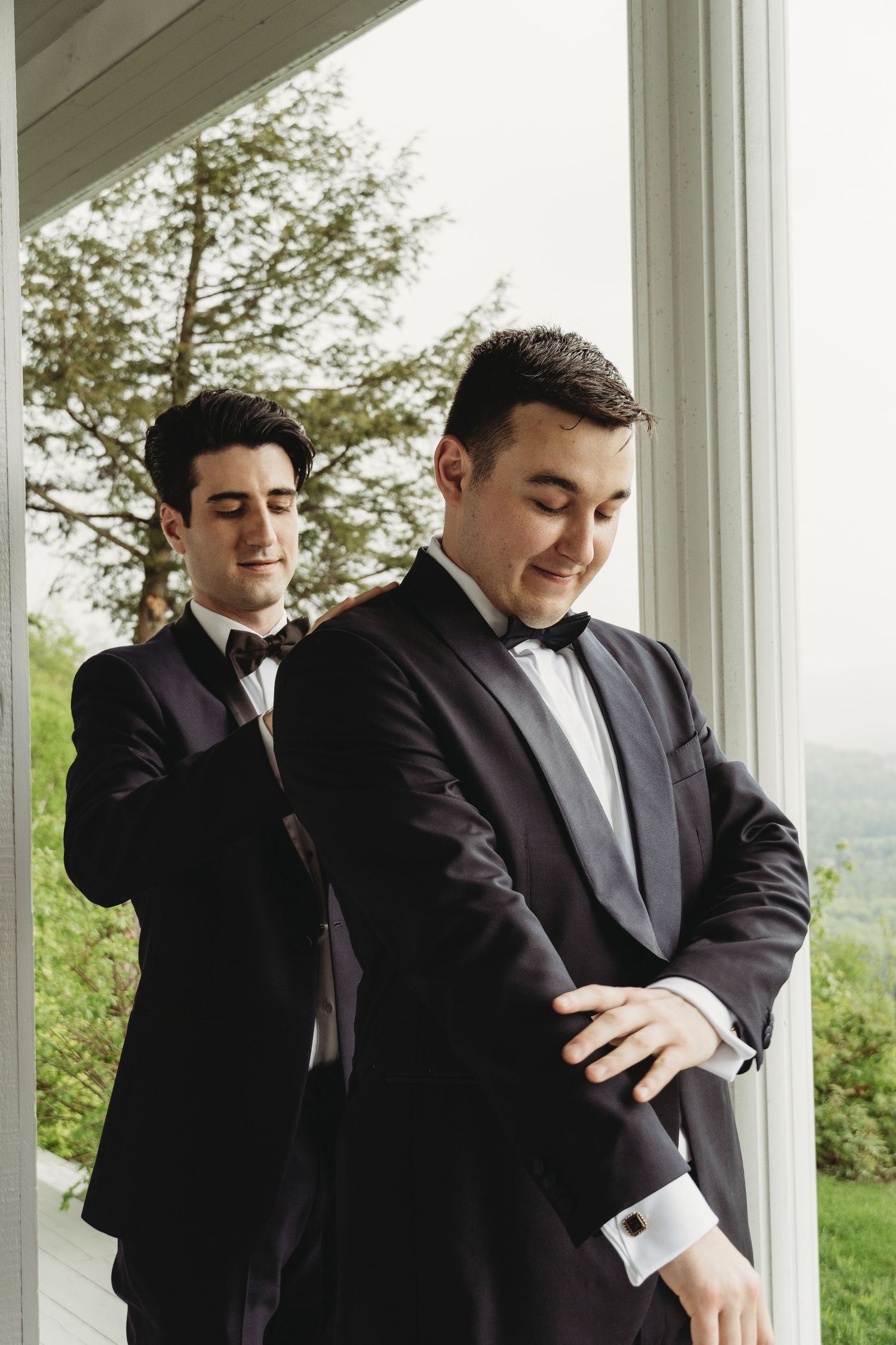 Two people in black tuxedos standing on a porch with a scenic view behind them.
