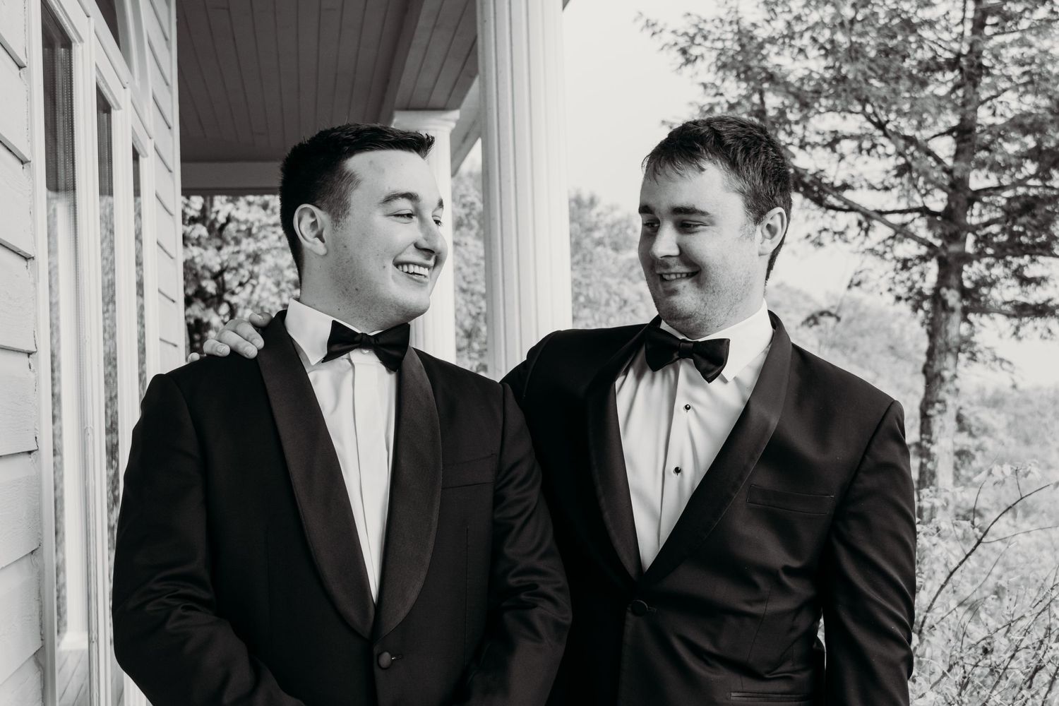 Two people in black bow ties and tuxedos sharing a candid moment on a covered porch.
