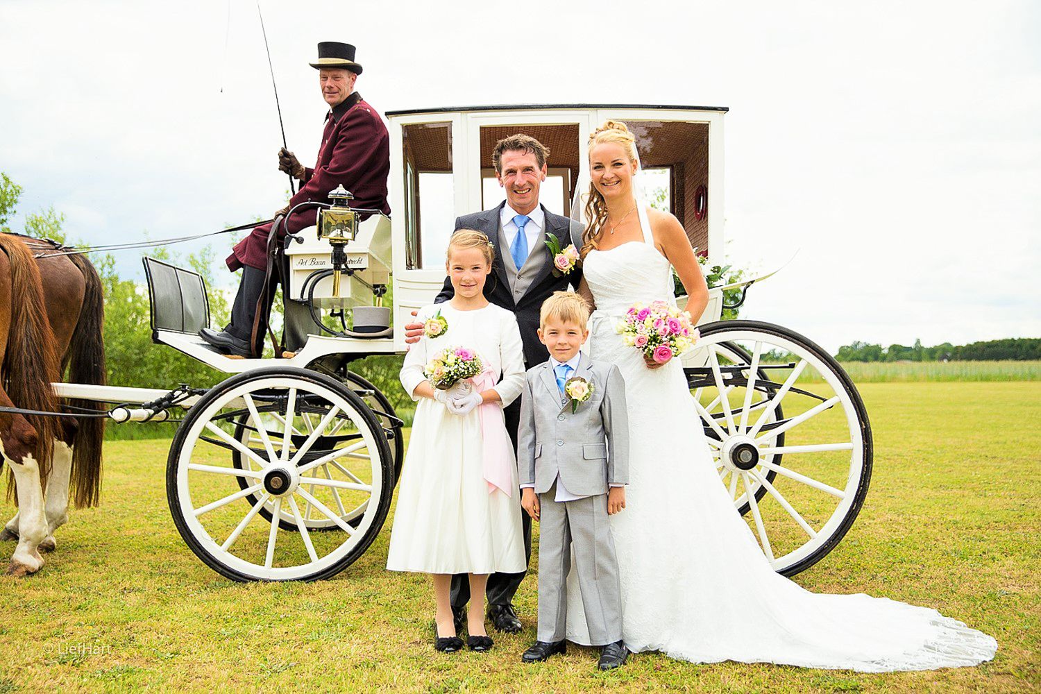 Classic wedding portrait with flower girls and carriage driver in front of white horse-drawn carriage.
