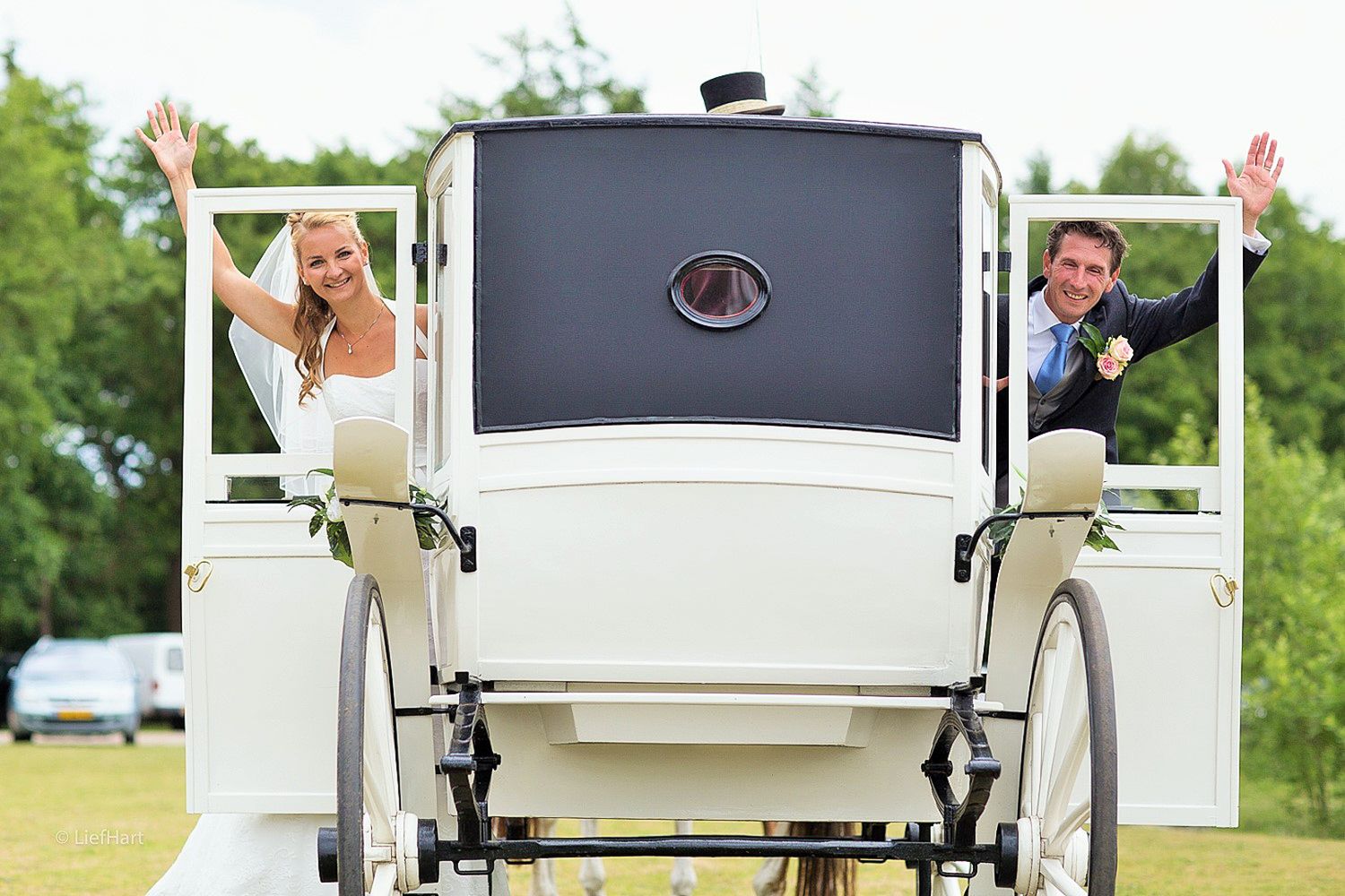 Happy newlyweds waving from the back of an elegant white horse-drawn carriage.
