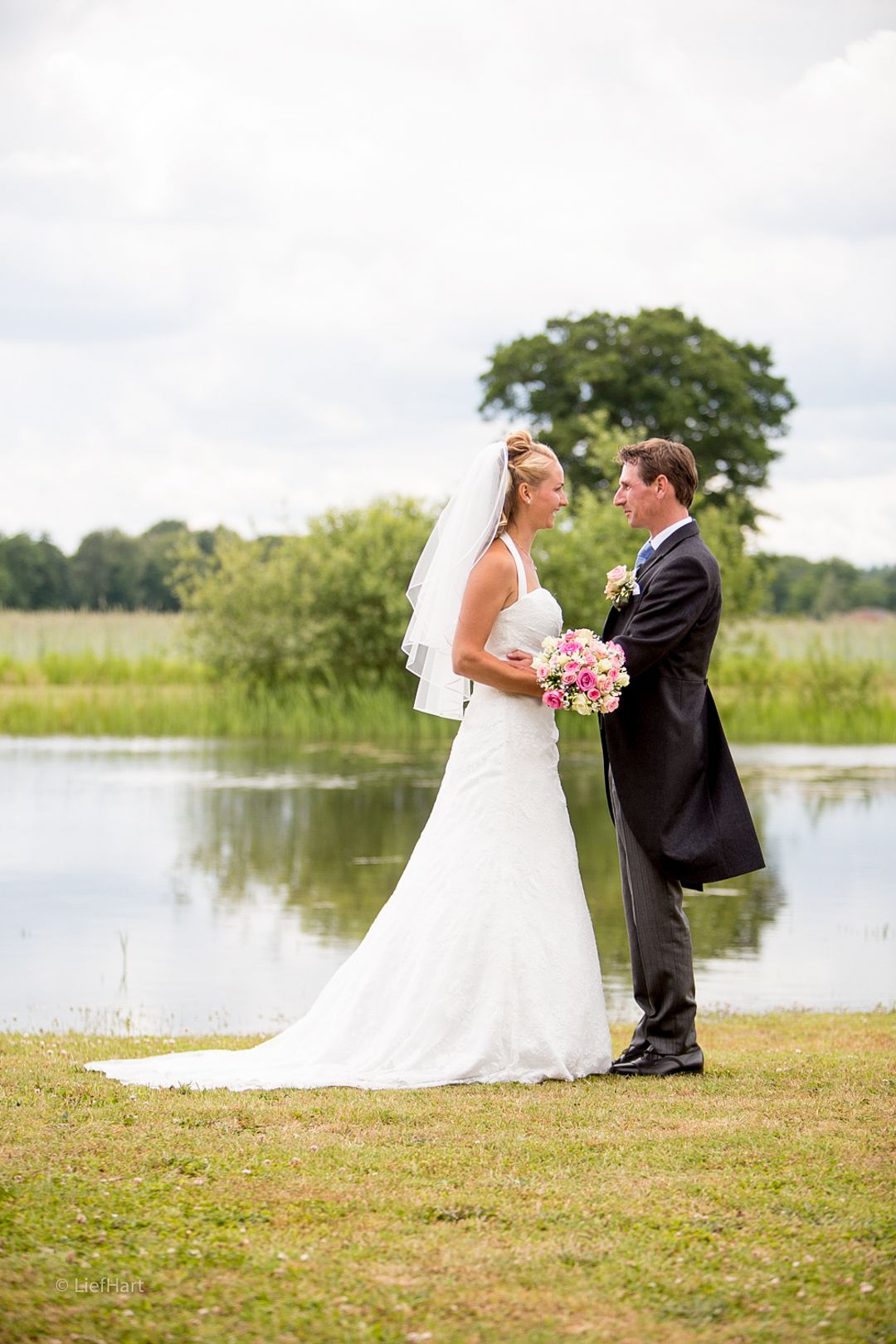 Romantic lakeside wedding portrait with couple standing on grassy shore.