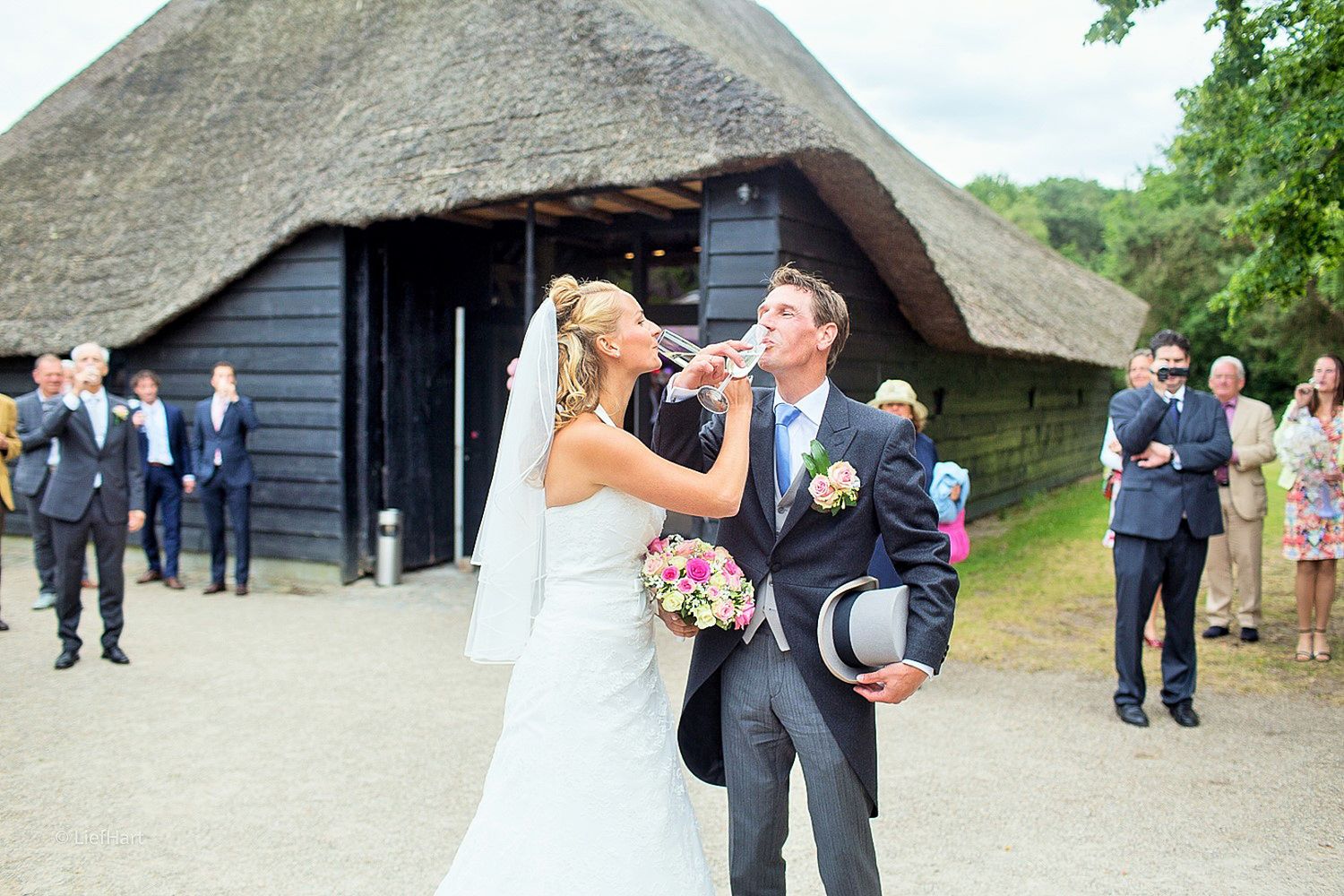 Joyful wedding moment captured outside rustic thatched-roof building with guests in background.