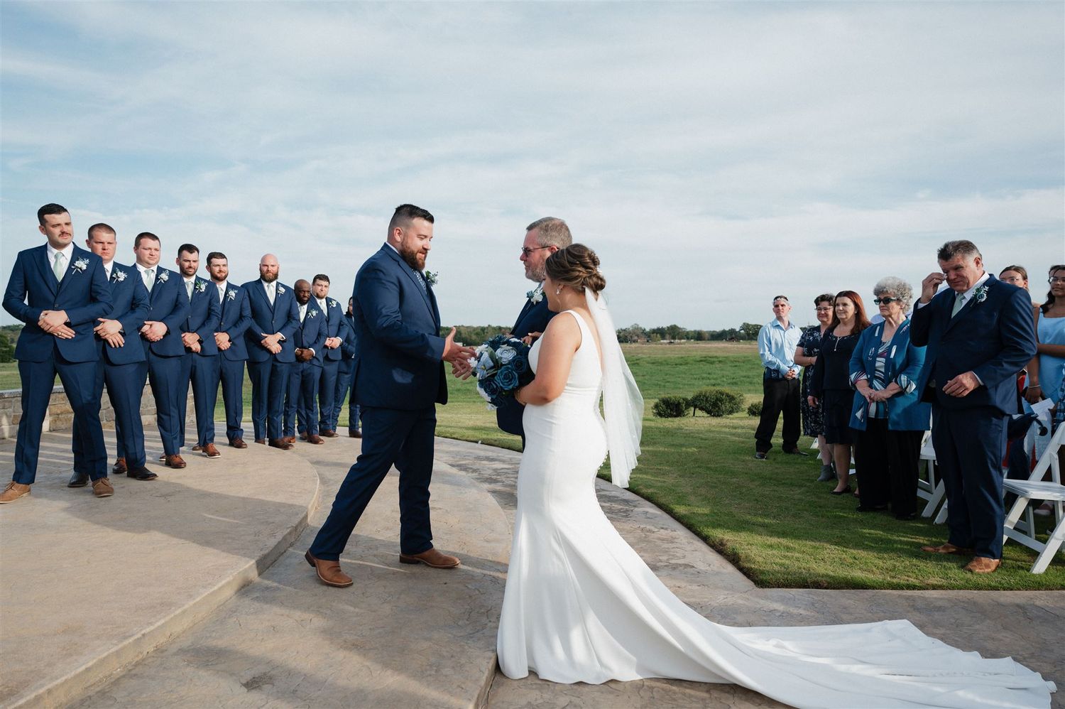 Couple sharing first dance moment during outdoor wedding with wedding party looking on.