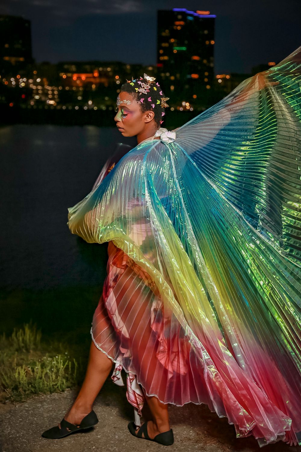 Figure in an iridescent rainbow cape dress poses against a city skyline backdrop at night.