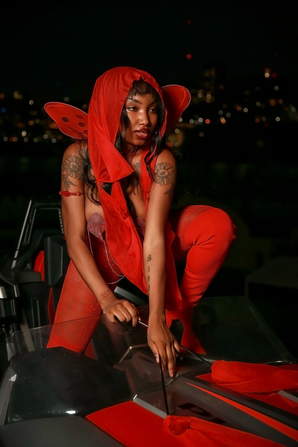 Person in vibrant red streetwear outfit poses dramatically at night on car hood with cityscape lights in background.