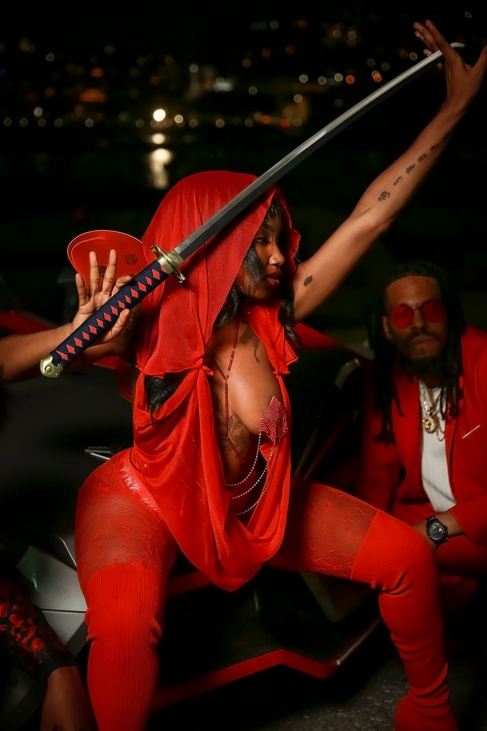 Performer in vibrant red outfit strikes dramatic pose with samurai sword against nighttime city backdrop.