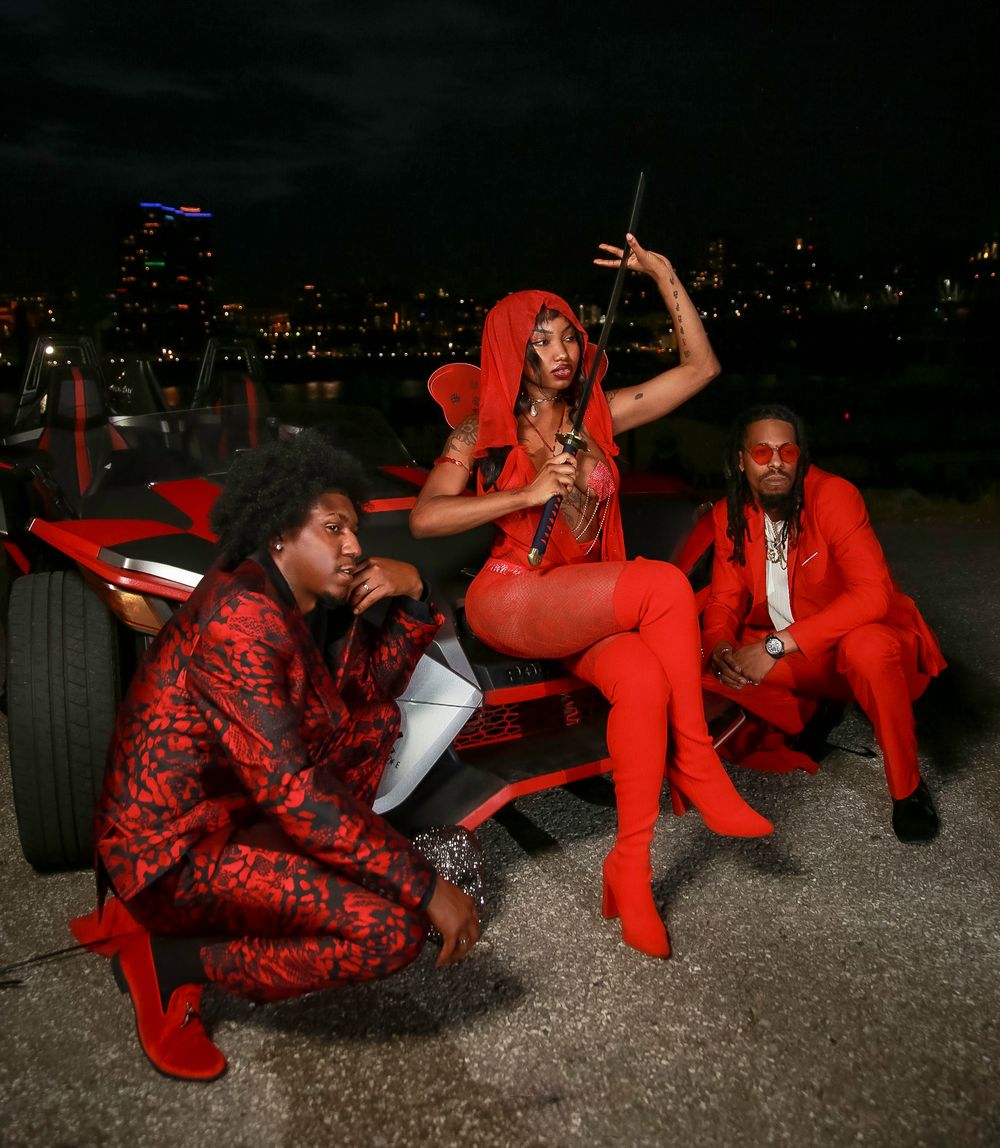 Three people in vibrant red outfits pose together at night with city lights visible in the background.