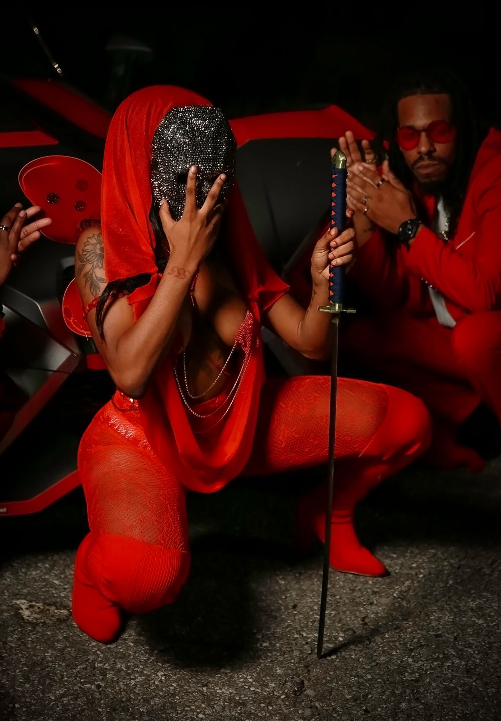 Performance art in red costumes with dramatic posing figures and beaded props against dark background.