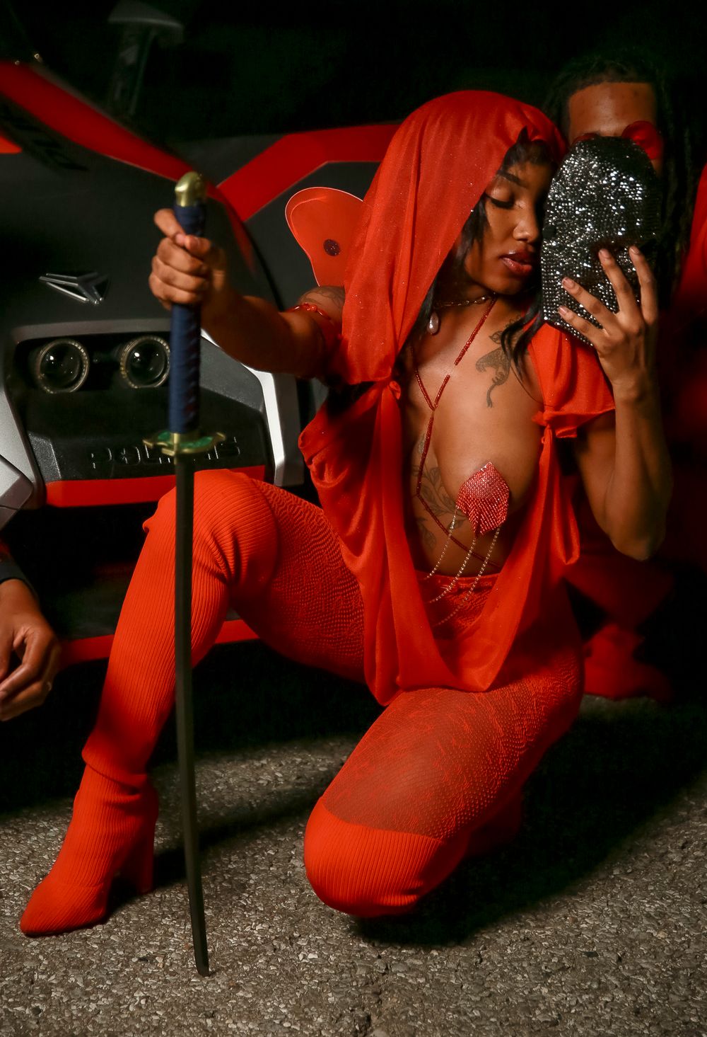 A person in a bright red hooded outfit poses with a phone and cane against a dark background.