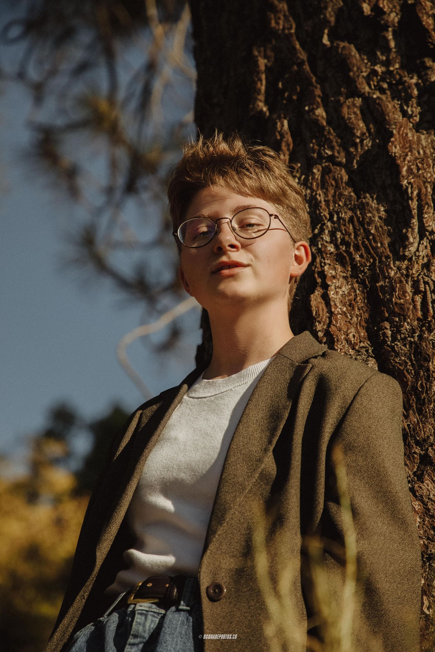 Casual portrait of person in glasses and brown blazer leaning against tree trunk.