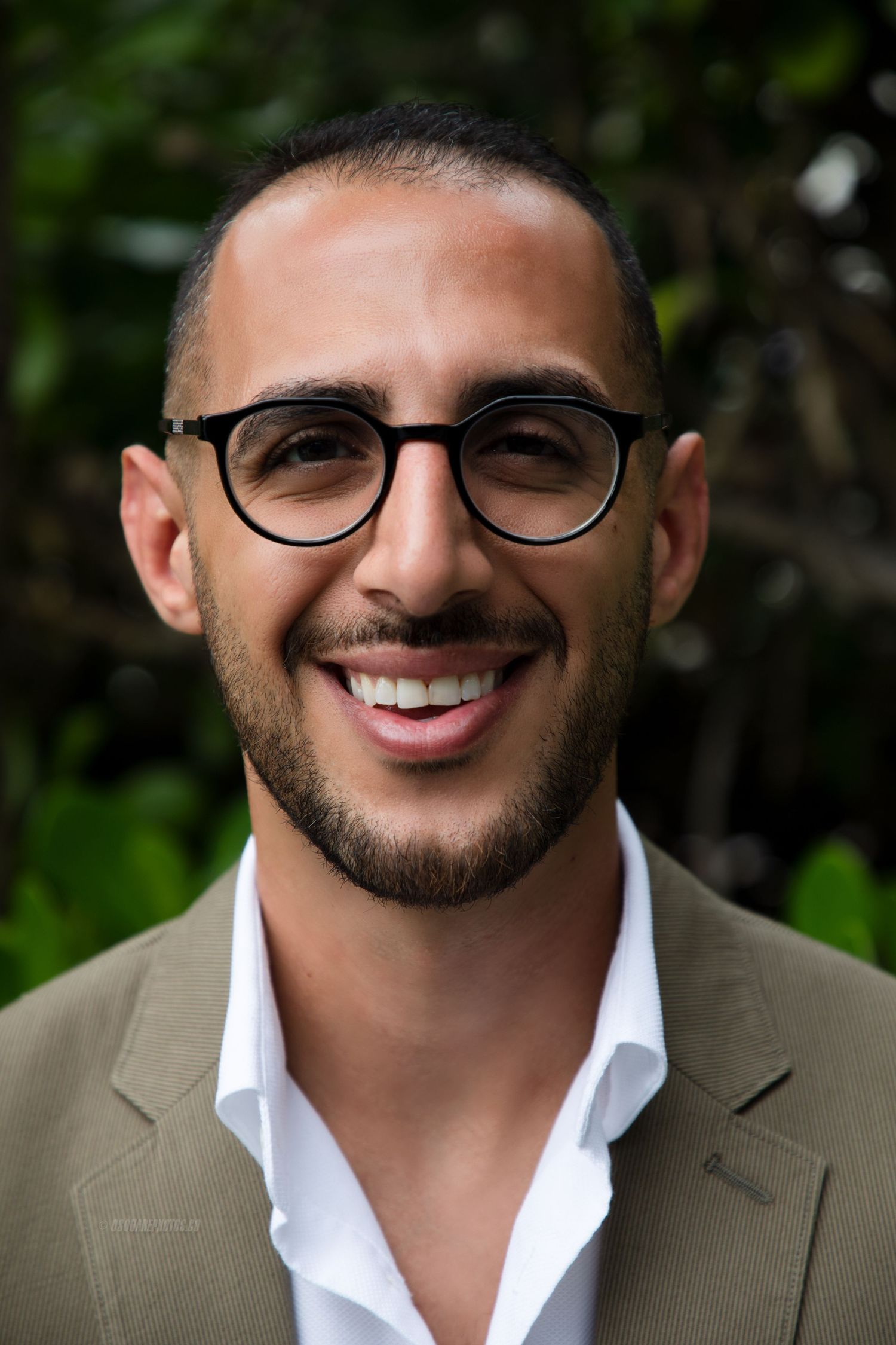 Professional headshot of person in glasses wearing tan blazer and white shirt against green foliage.