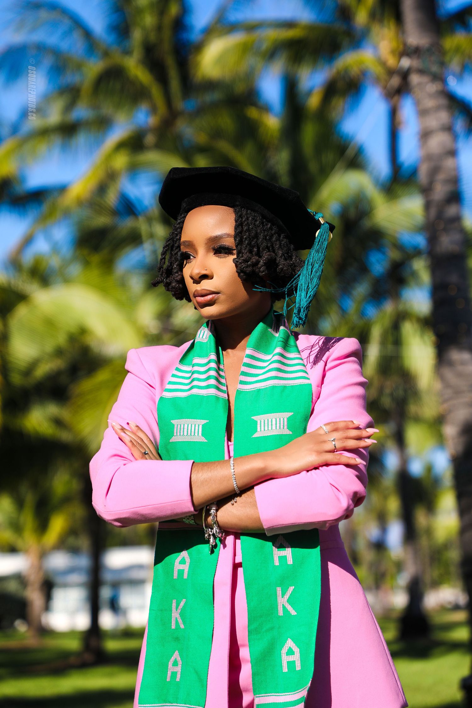 Graduate in pink and green academic regalia poses confidently with palm trees in background.