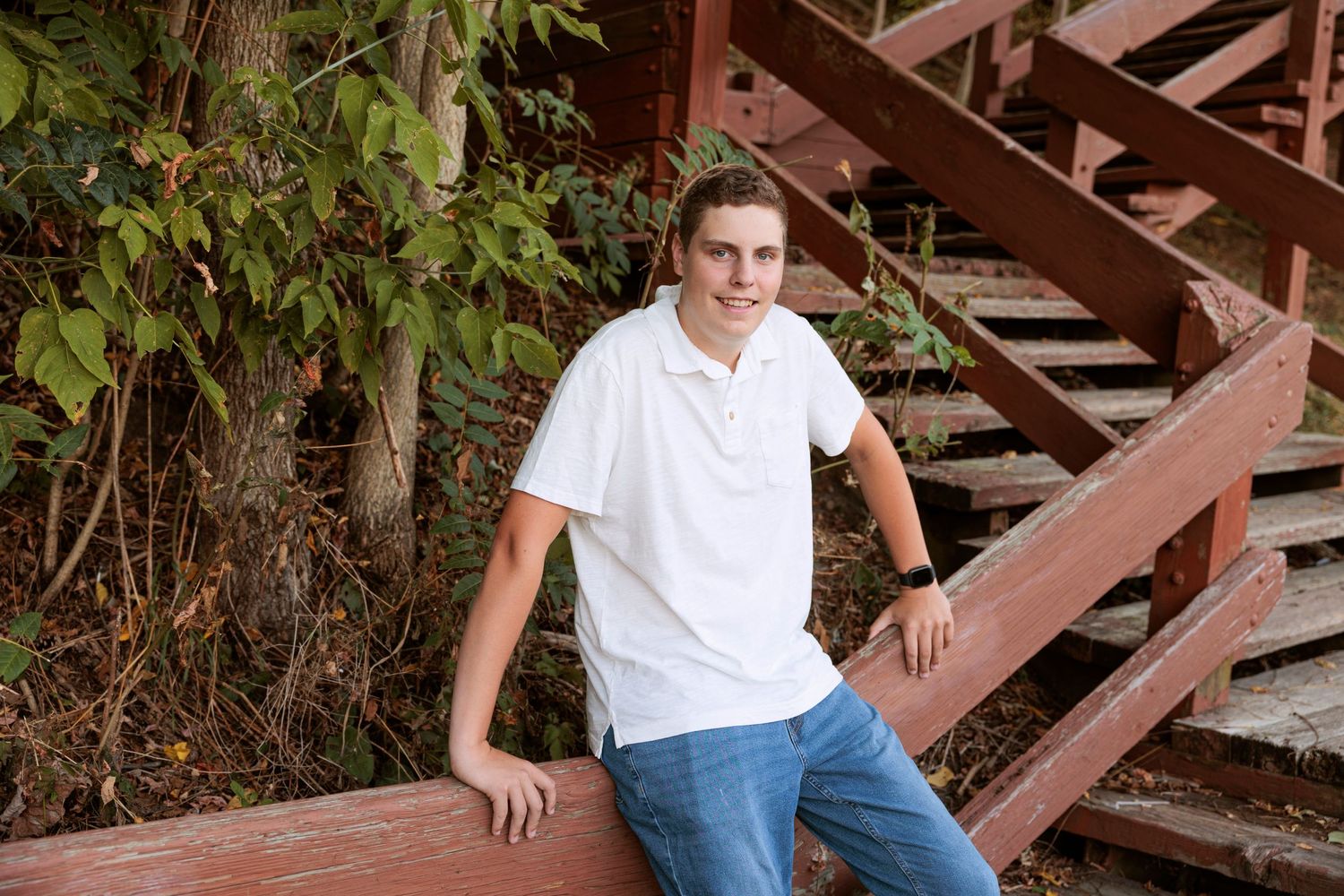 A casual portrait of someone posing on wooden stairs while wearing a white polo and blue jeans.