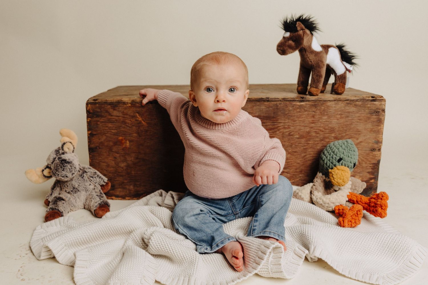 Baby in jeans sits against wooden box with stuffed animals.