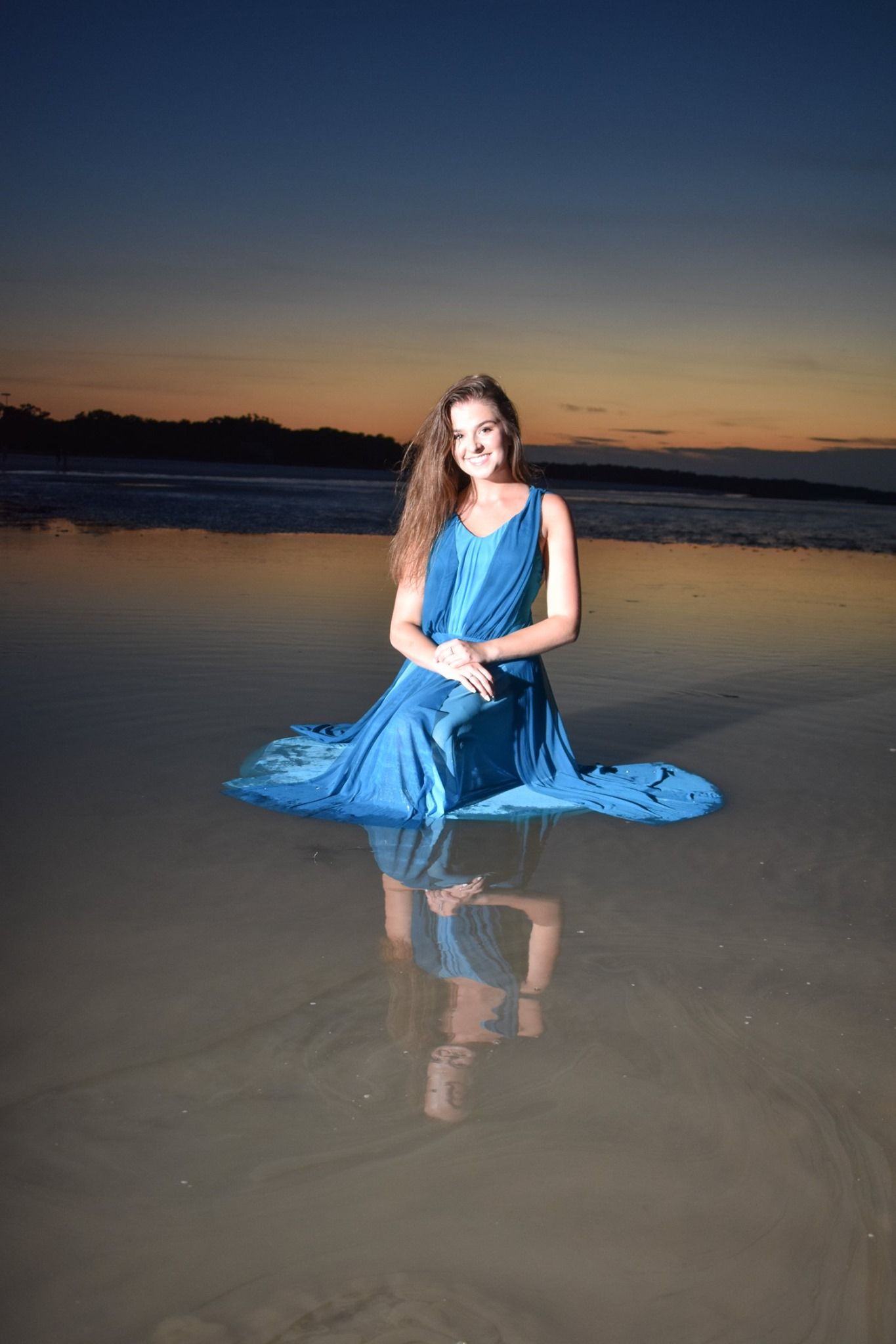 Young woman in flowing blue gown sitting in shallow ocean water during colorful twilight sunset.