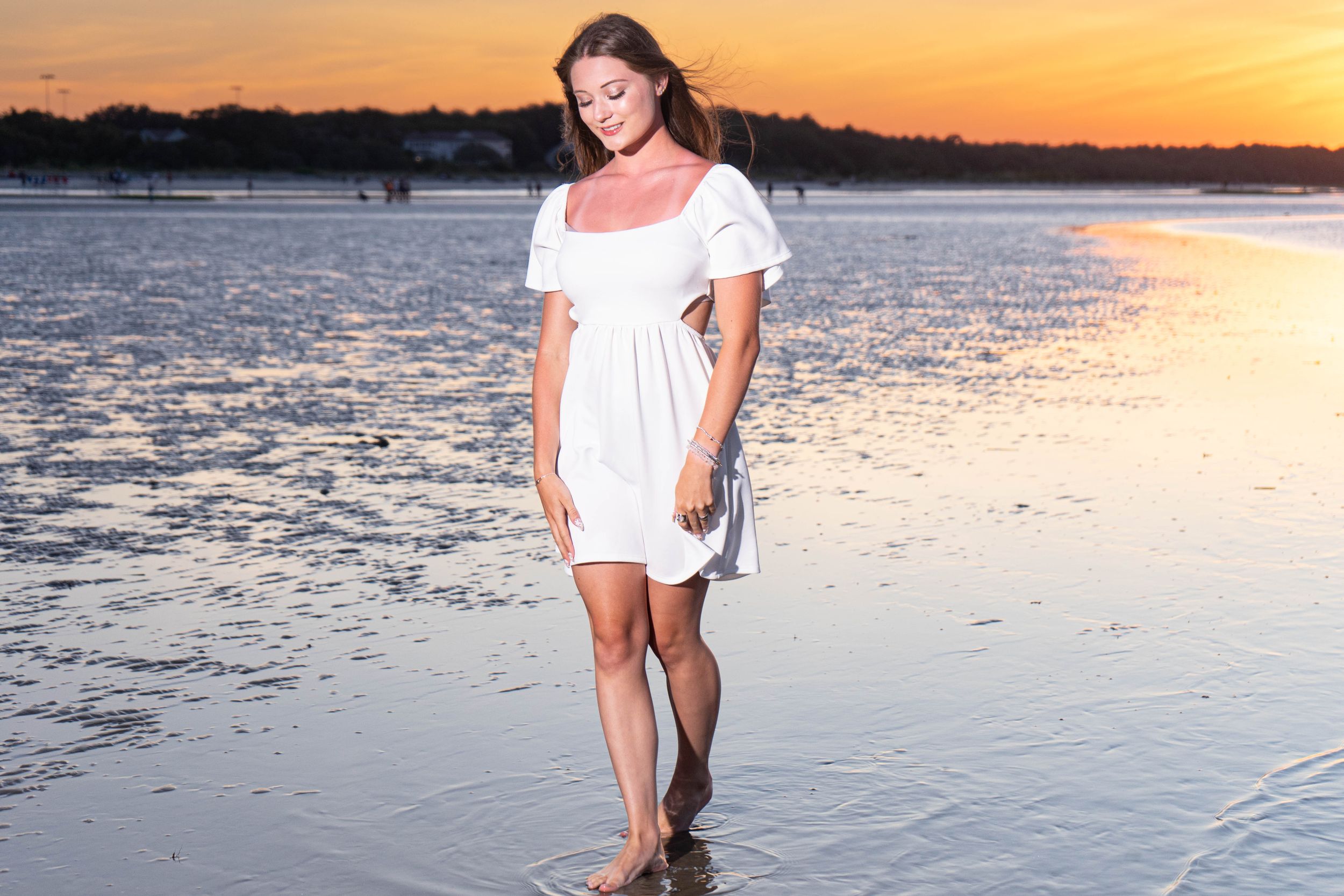 Young woman in white dress walking on wet beach shore during stunning orange sunset reflection.