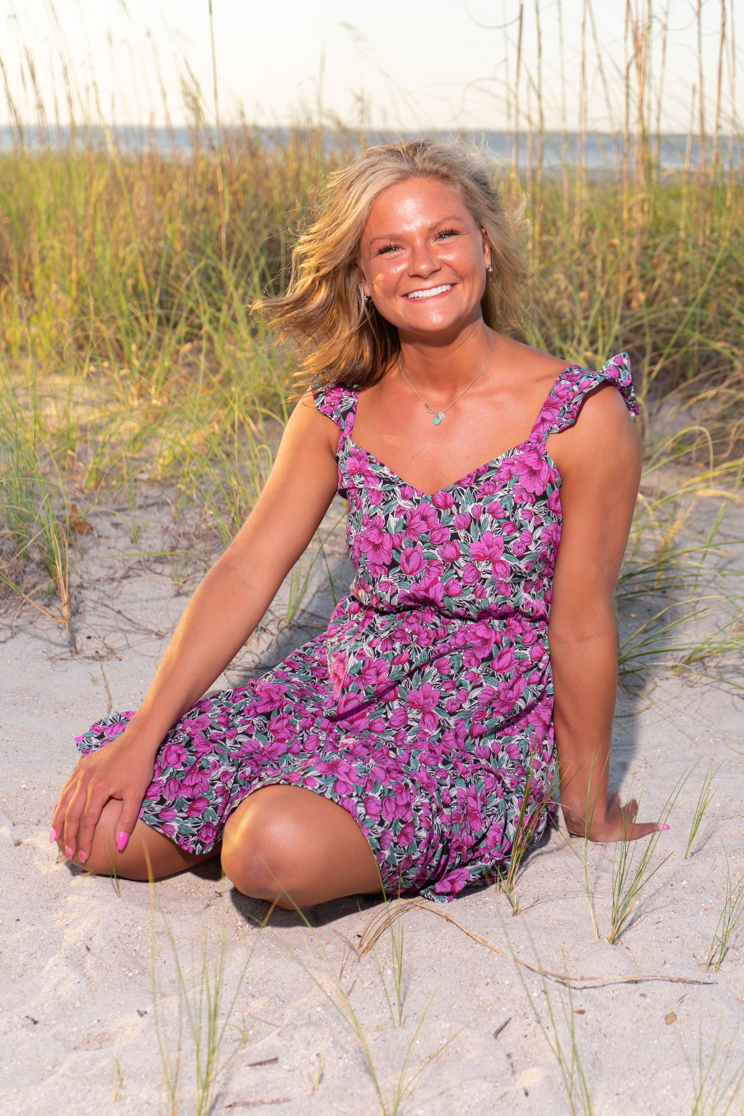 Smiling blonde woman in pink floral dress sitting on beach sand dunes with sea oats in background.