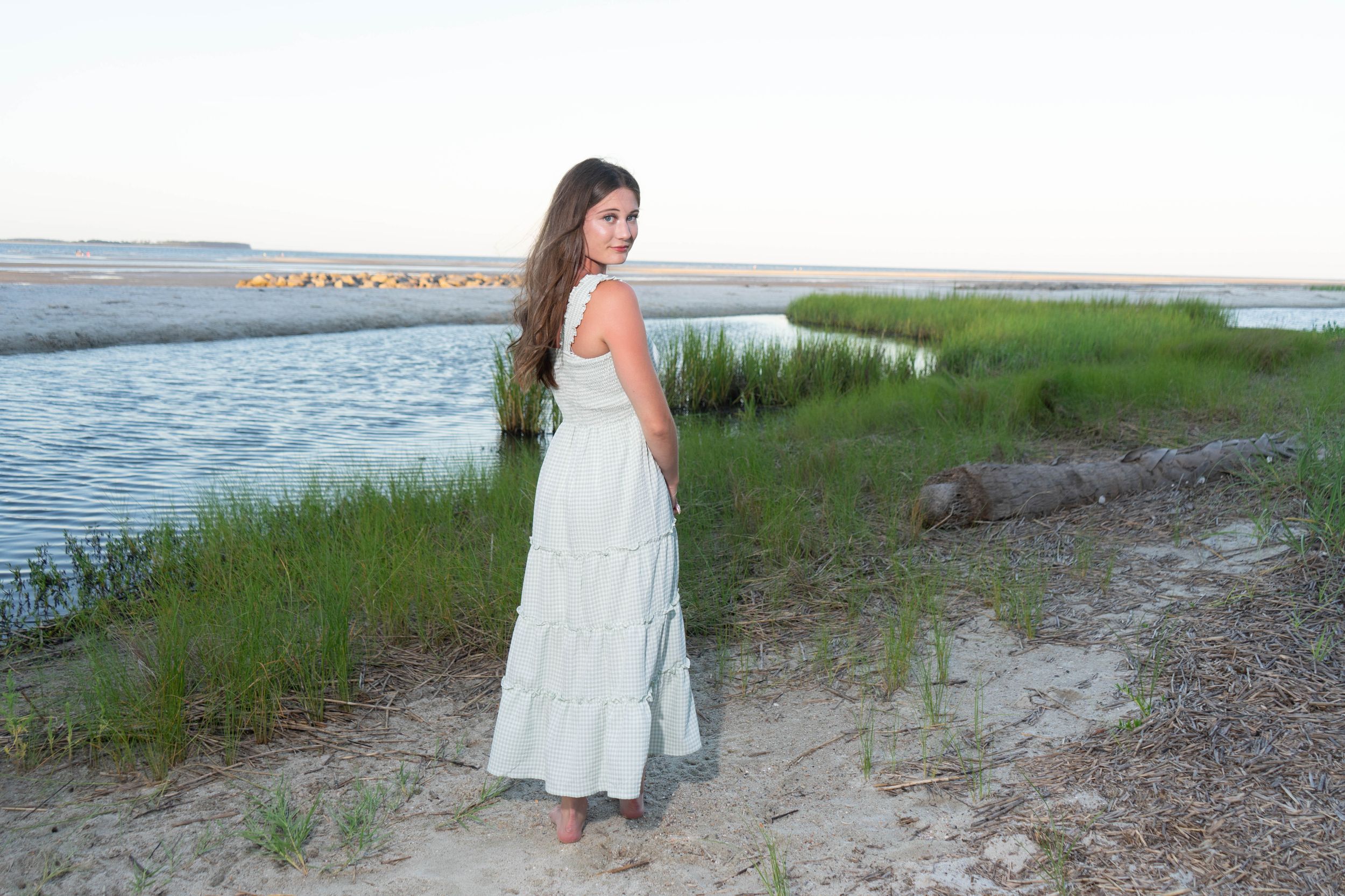 Young woman in white maxi dress standing on sandy beach shore with marsh grass and calm water behind her.