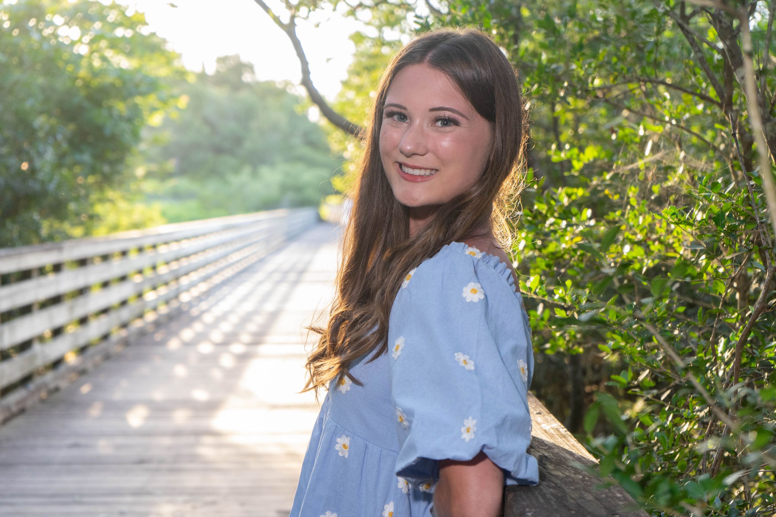 Smiling young woman in blue floral dress posing on wooden boardwalk surrounded by lush green trees.