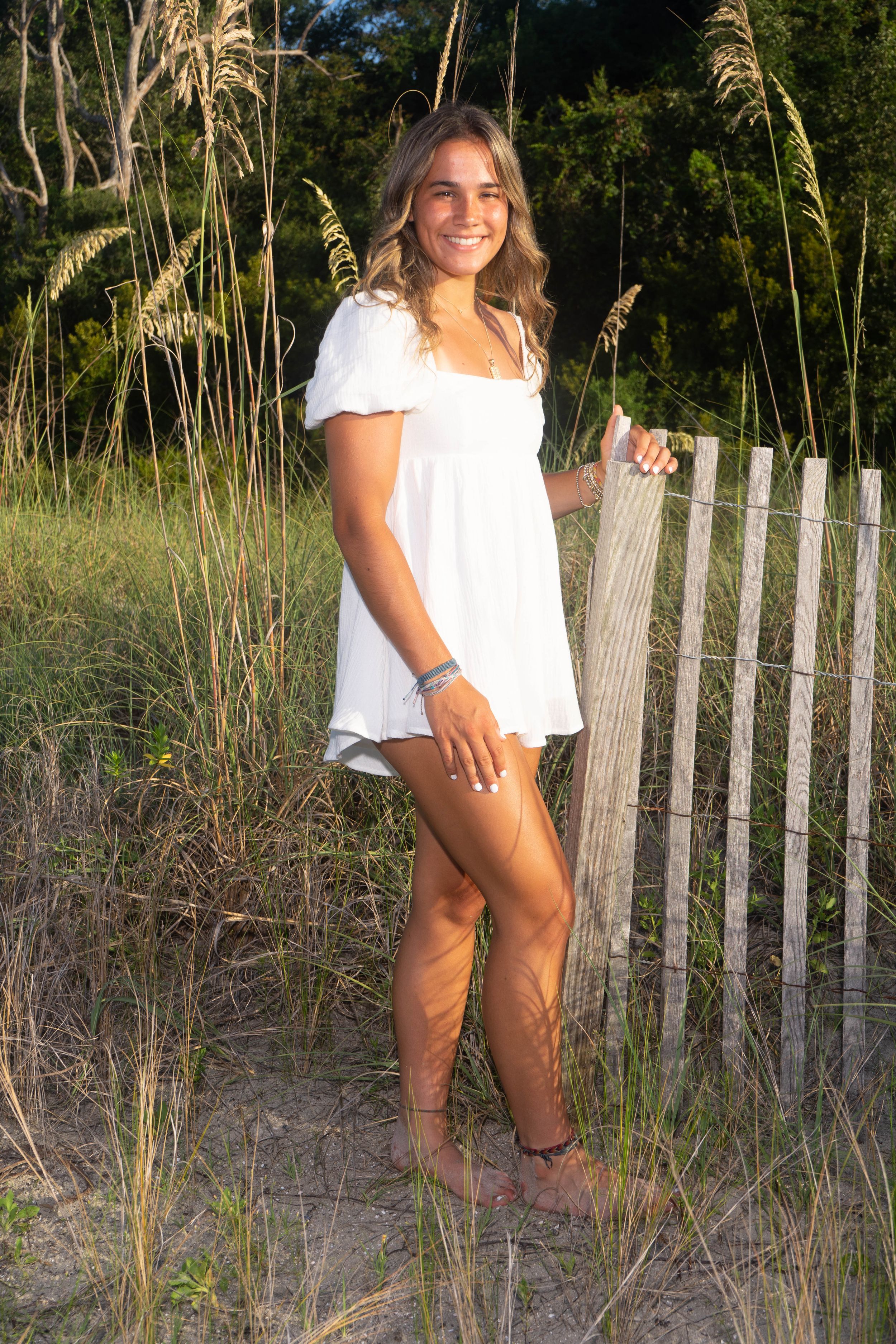 Smiling young woman in white mini dress standing by wooden fence among tall sea oats at beach.