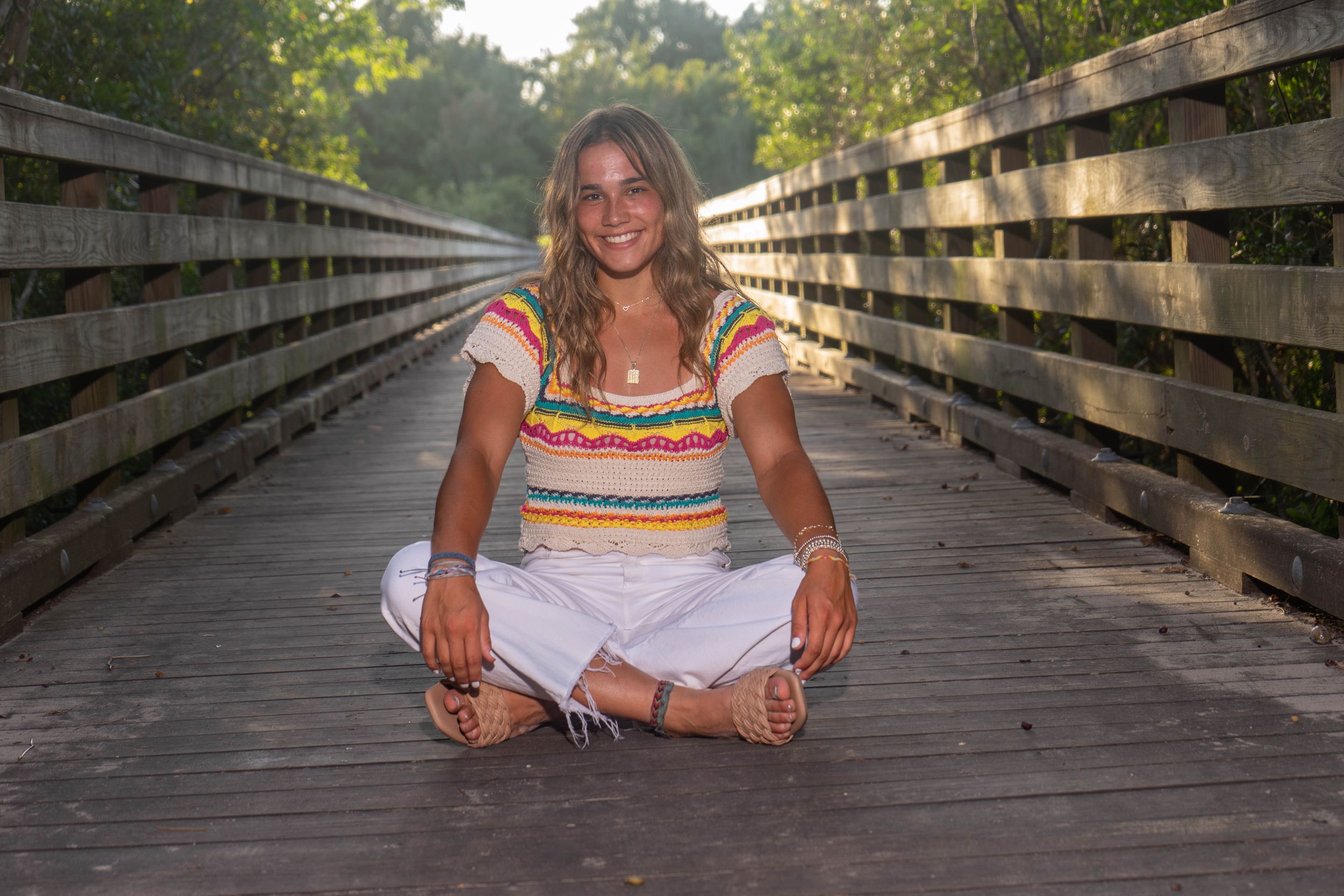 Smiling young woman in colorful top and white pants sitting cross-legged on wooden boardwalk bridge.