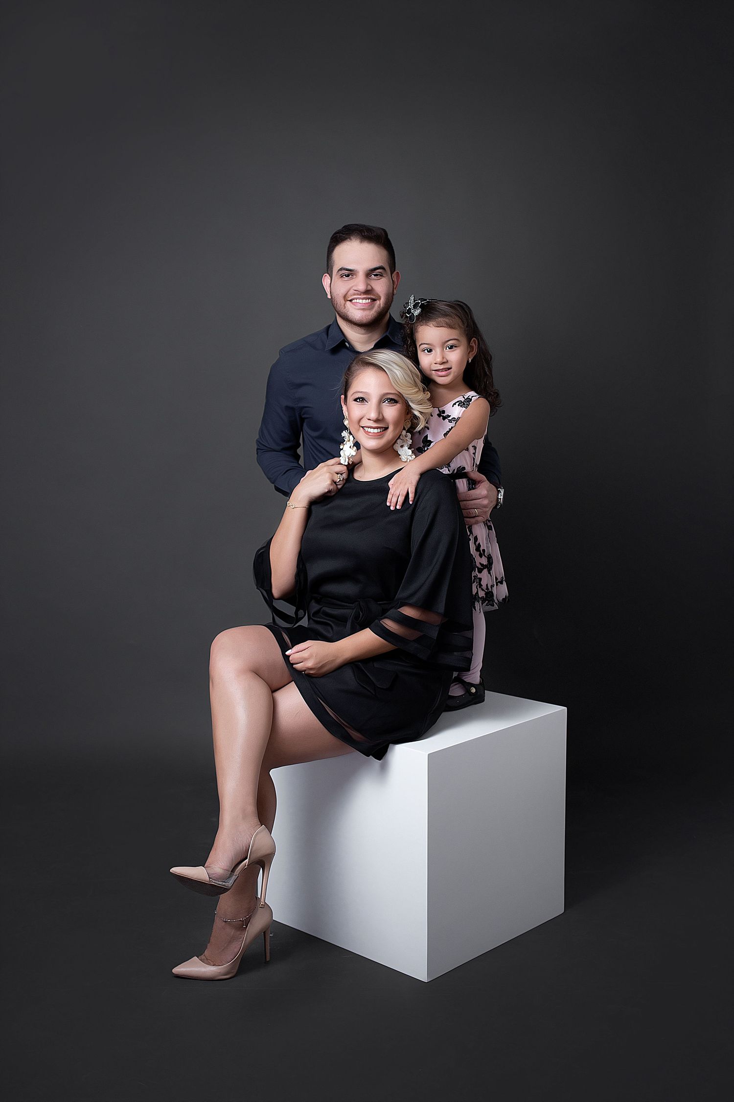 Studio portrait session showing playful poses of family on white cube against dark background.