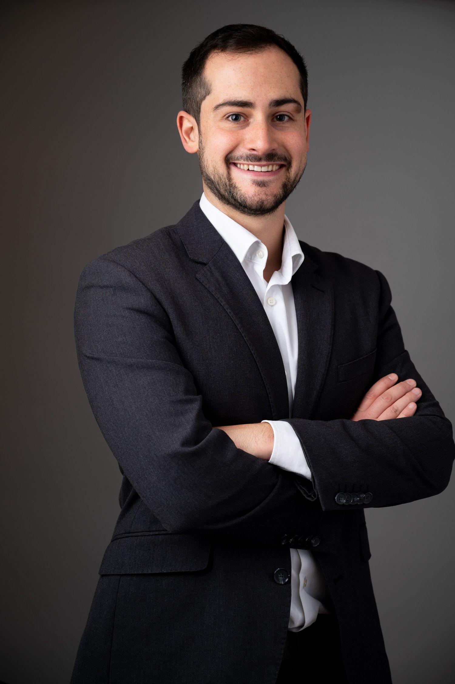 Professional headshot of a businessman in a dark suit jacket and white shirt against a grey background.