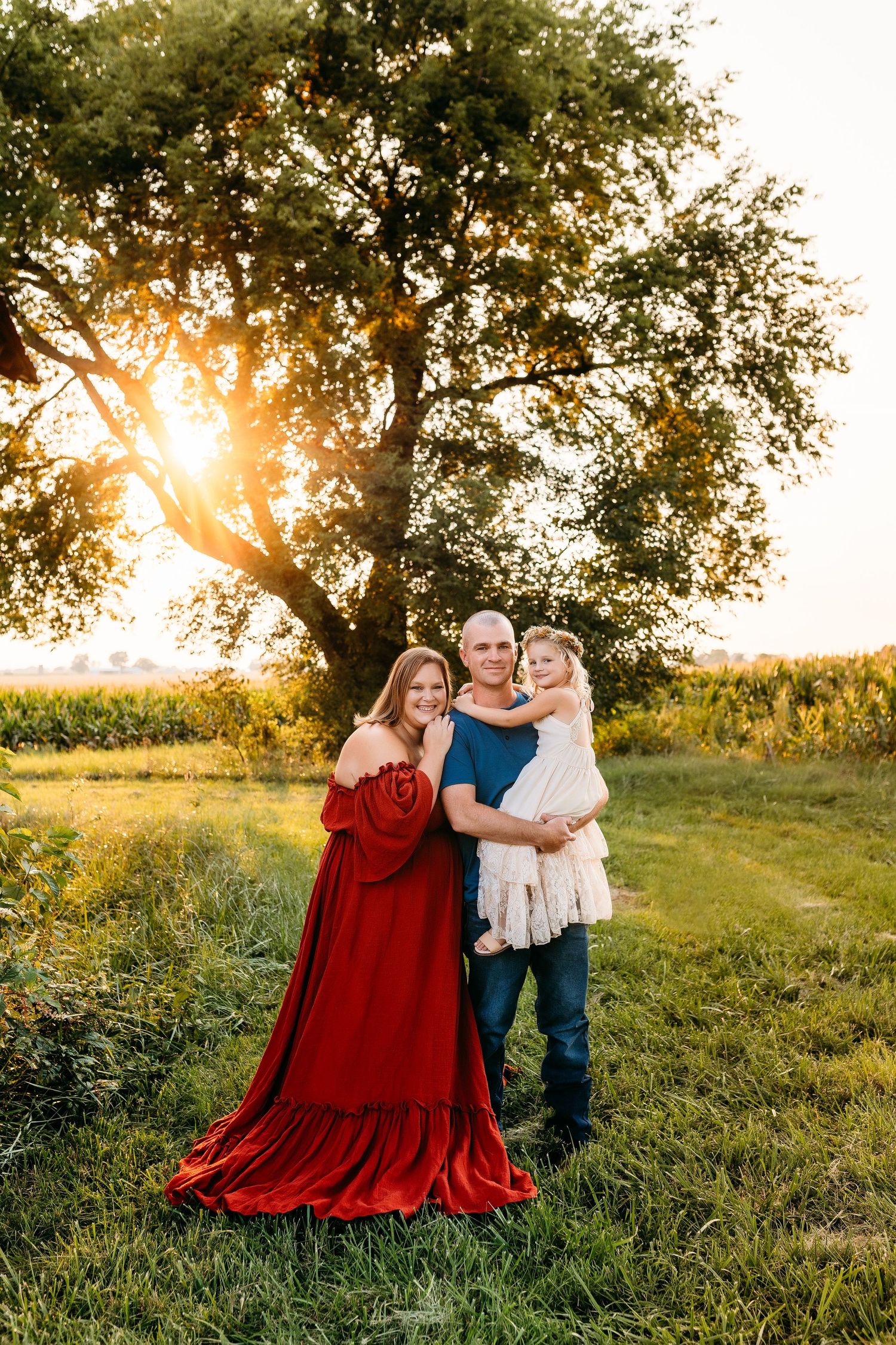 A family shares a tender moment at golden hour beside a large tree and red barn.