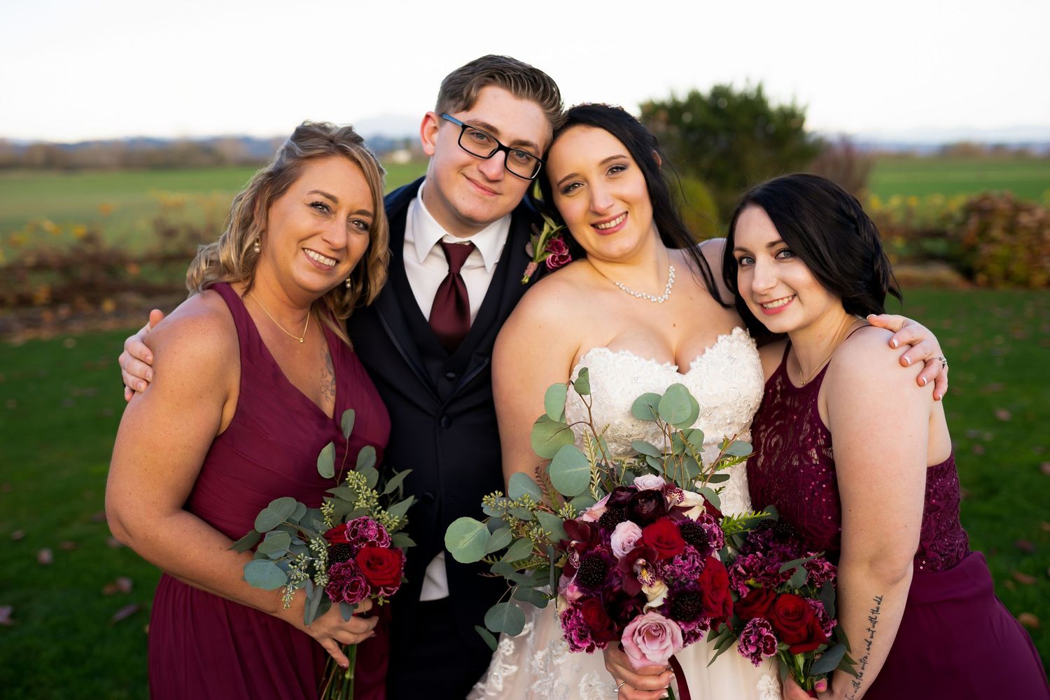 A wedding party in burgundy dresses and formal attire poses together outdoors.